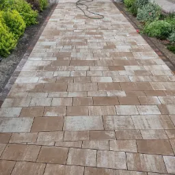 Ułożona aleja z dwukolorowej kostki brukowej prowadząca do domu, obrzeża z ciemniejszej kostki, po bokach rabaty z zielenią, wąż ogrodowy leży na kostce.