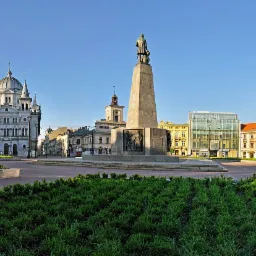 Panorama miejskiego placu z pomnikiem, kościołem z kopułą i różnorodną architekturą w słoneczny dzień.