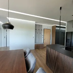 Nowoczesne wnętrze domu z drewnianą podłogą, stołem, krzesłami, wyspą kuchenną z pionowymi listwami, wbudowaną ławką z wieszakami i oświetleniem LED w suficie.