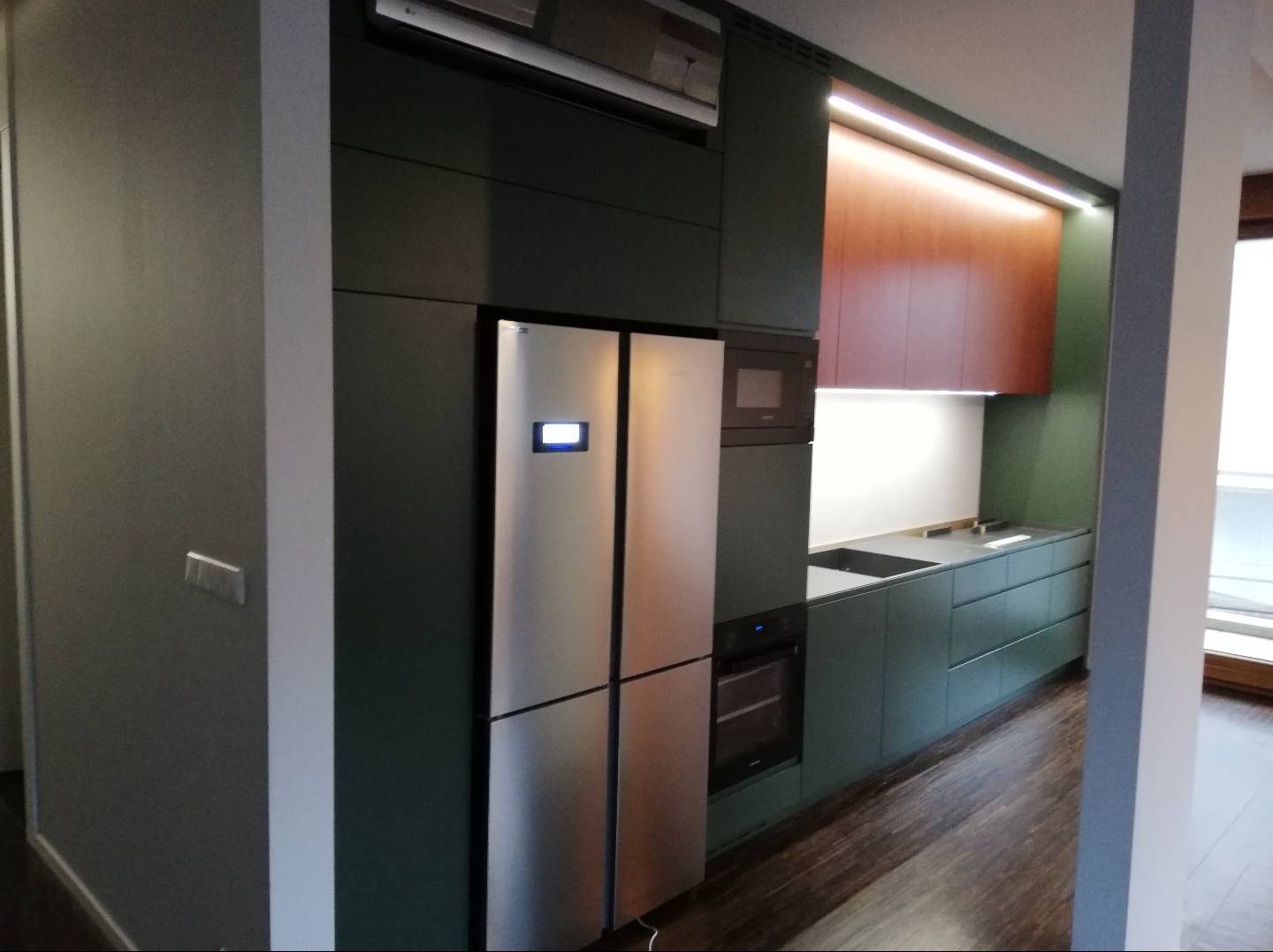 Nowoczesna kuchnia z zabudową w odcieniach zieleni i brązu, z lodówką side-by-side i oświetleniem LED podszafkowym, widok z boku.