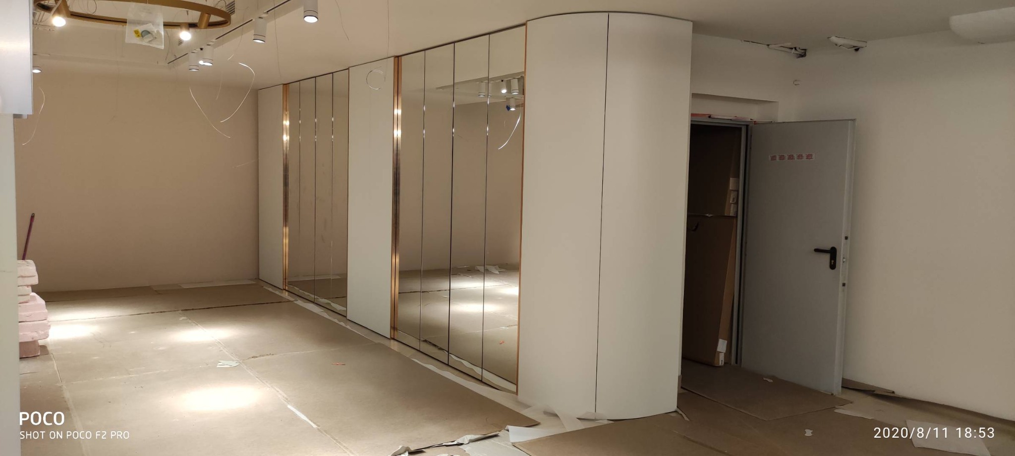 Wnętrze w trakcie wykańczania z zamontowaną dużą szafą wnękową z lustrzanymi drzwiami i złotymi obramowaniami, widoczne niedokończone podłogi i instalacje sufitowe.