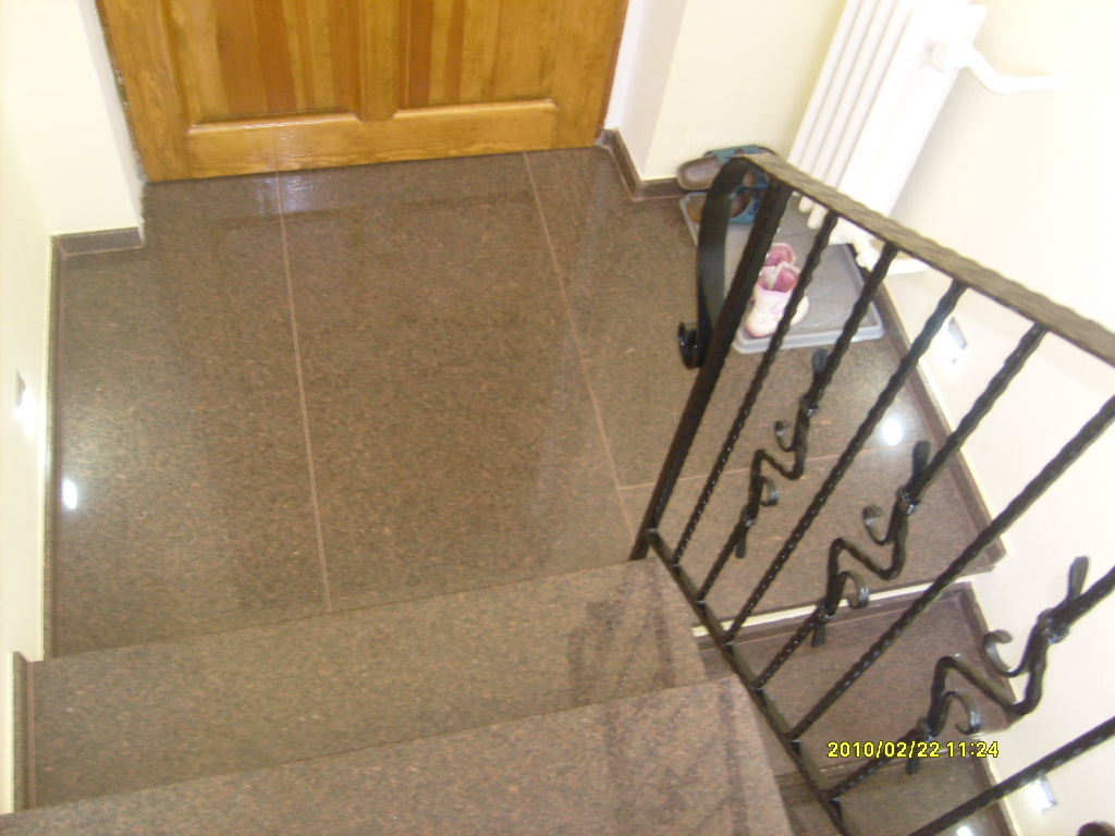 Granitowe schody wewnętrzne z czarną metalową balustradą, widok z góry, przy drewnianych drzwiach wejściowych, z widocznym fragmentem grzejnika i obuwiem w tle.