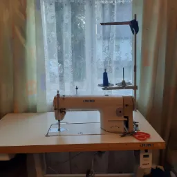 Maszyna do szycia JUKI na stole krawieckim, z niebieską nicią na szpulce, nożyczkami z czerwonymi uchwytami i zasłoniętym oknem w tle.