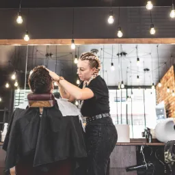 Klient w trakcie strzyżenia w nowoczesnym salonie z ceglaną ścianą i oświetleniem żarówkowym, fryzjerka w czarnym ubraniu z warkoczami przy pracy.