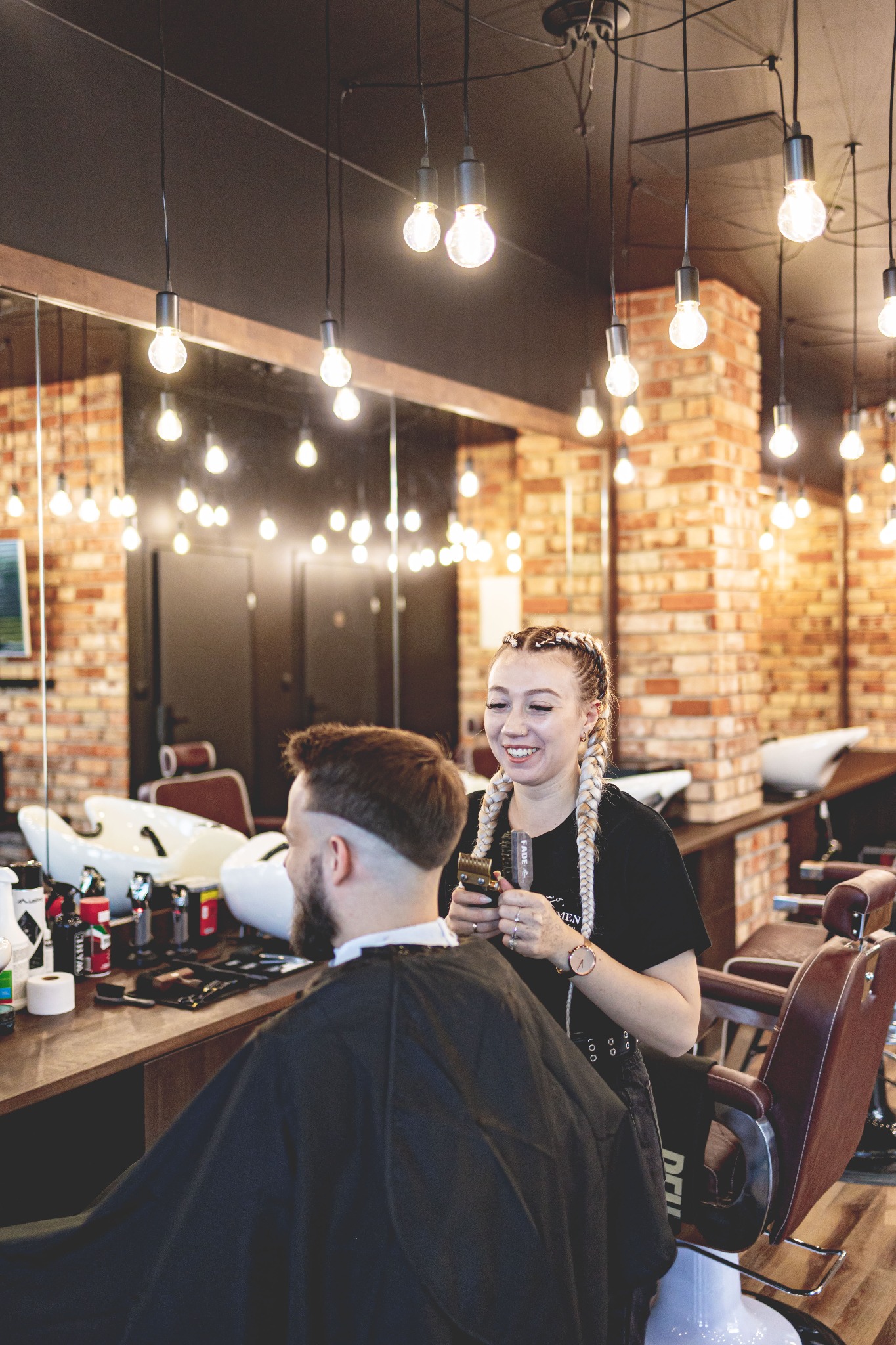 Uśmiechnięta blondynka z warkoczami, ubrana na czarno, trzyma maszynkę do strzyżenia w dłoniach, przygotowując się do strzyżenia brodatego mężczyzny w pelerynie w eleganckim salonie z ceglanymi...