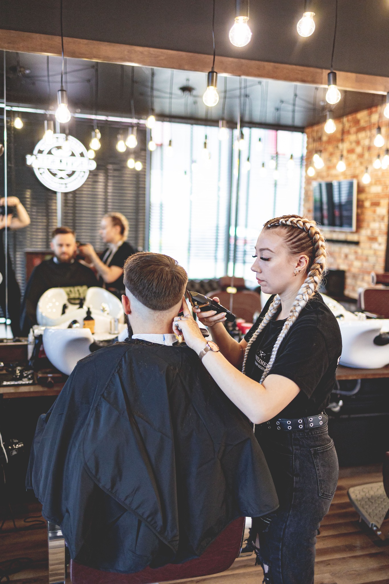 Fryzjerka z blond warkoczami strzyże klienta maszynką w salonie z ceglaną ścianą i lustrami.