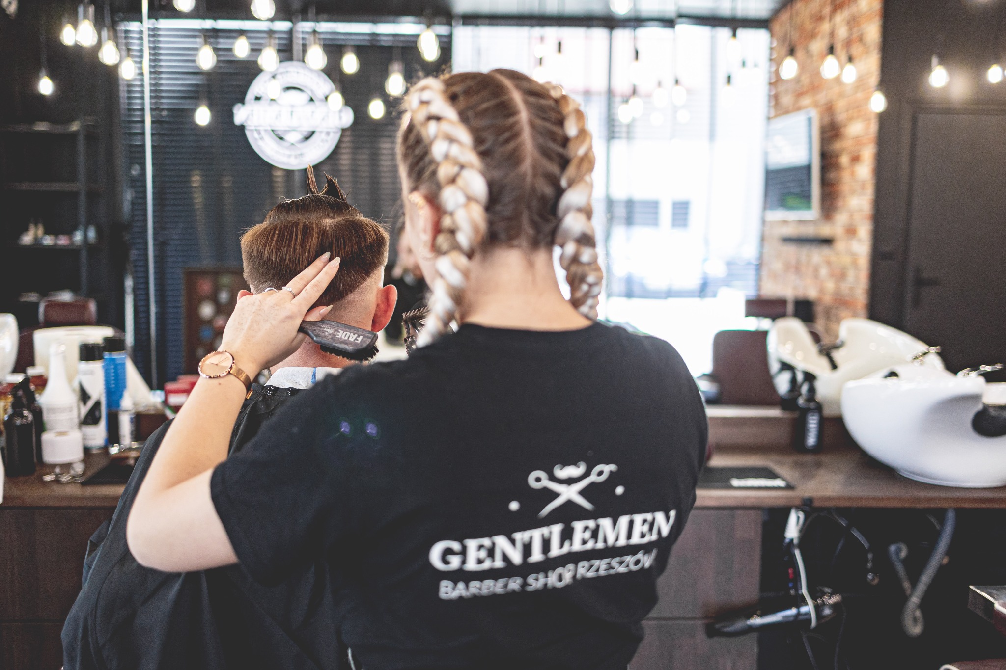 Strzyżenie męskie w salonie: fryzjerka z blond warkoczami modeluje włosy klientowi przy użyciu grzebienia i maszynki FADE, widoczne logo salonu 'Gentlemen Barber Shop Rzeszów' na koszulce.