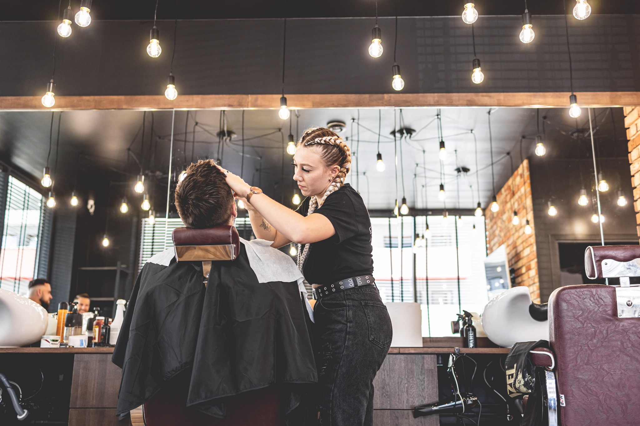 Klient w trakcie strzyżenia w nowoczesnym salonie z ceglaną ścianą i oświetleniem żarówkowym, fryzjerka w czarnym ubraniu z warkoczami przy pracy.