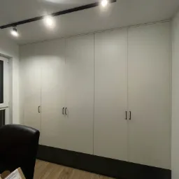 Wysoka szafa wnękowa z jasnymi frontami i ciemnym cokołem, oświetlenie szynowe na suficie, fragment biurka z kartonem w dolnym lewym rogu.