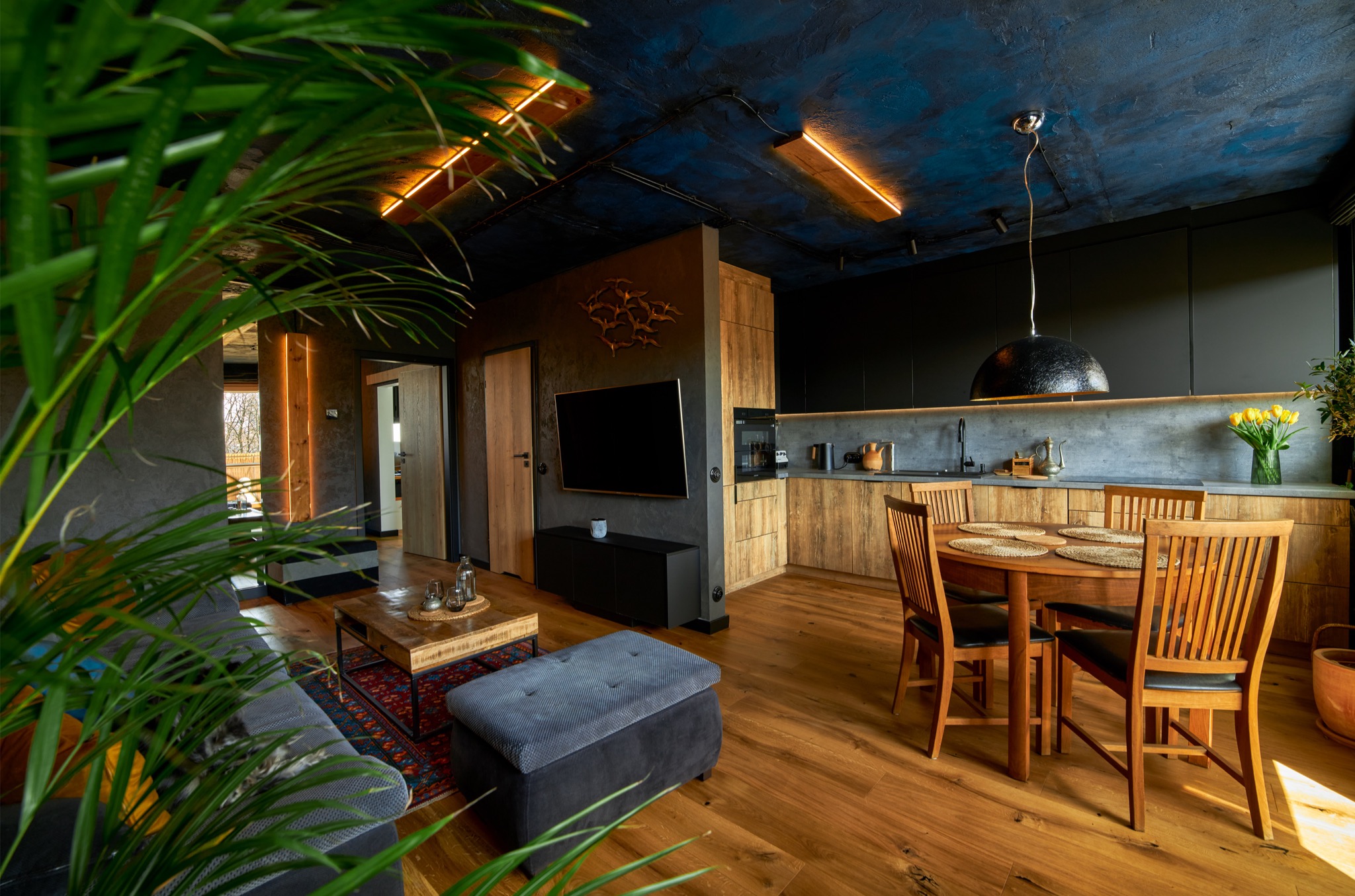 Wnętrze domu z otwartą kuchnią i salonem, drewniane meble kuchenne z czarnymi frontami, okrągły stół z drewnianymi krzesłami, sufit w odcieniach niebieskiego z podłużnymi lampami, podłoga z desek.