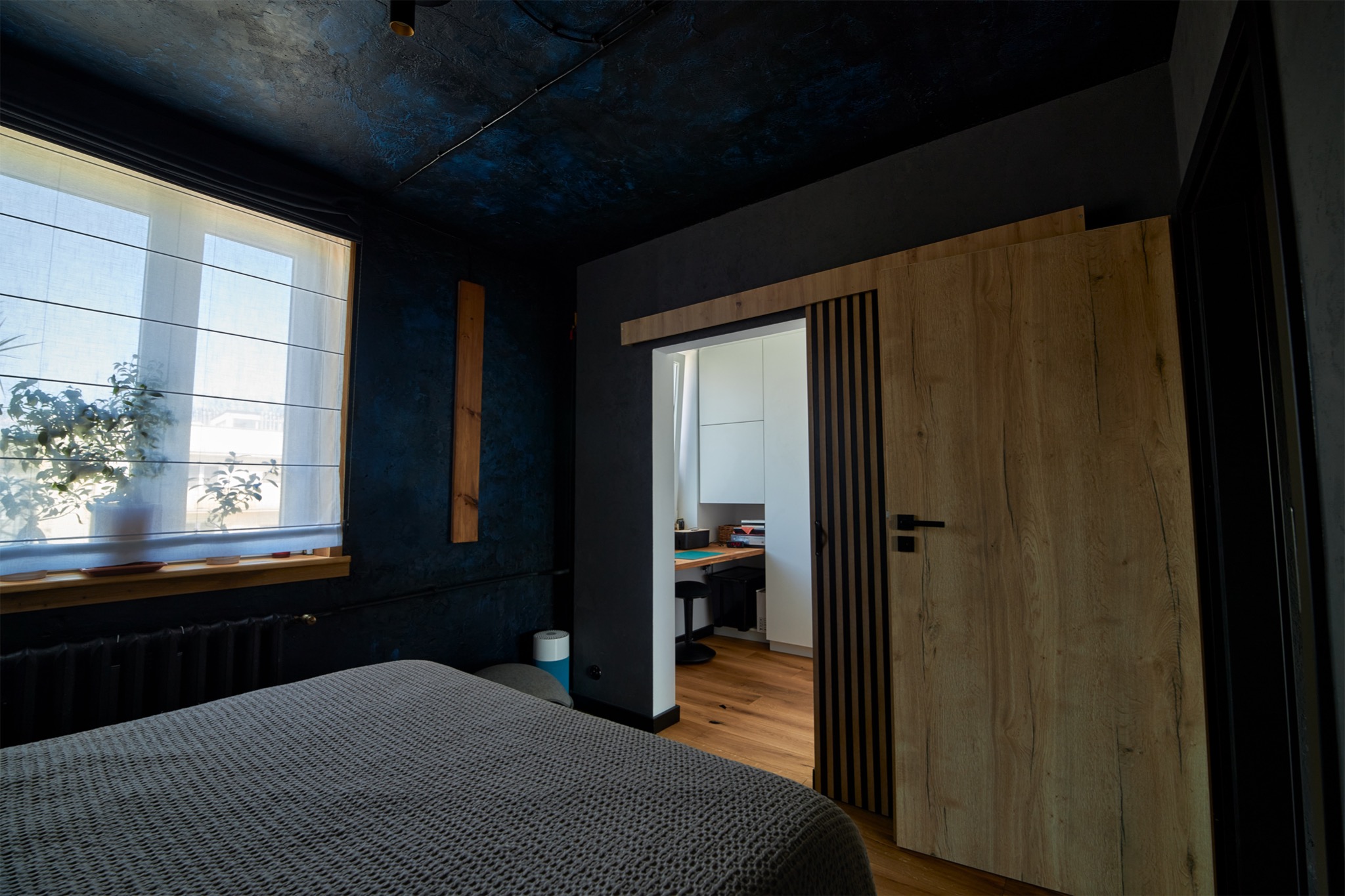 Nowoczesna sypialnia z ciemnymi ścianami, drewnianym panelem i przesuwnymi drzwiami z okleiną drewnopodobną prowadzącymi do jasnego gabinetu z biurkiem.