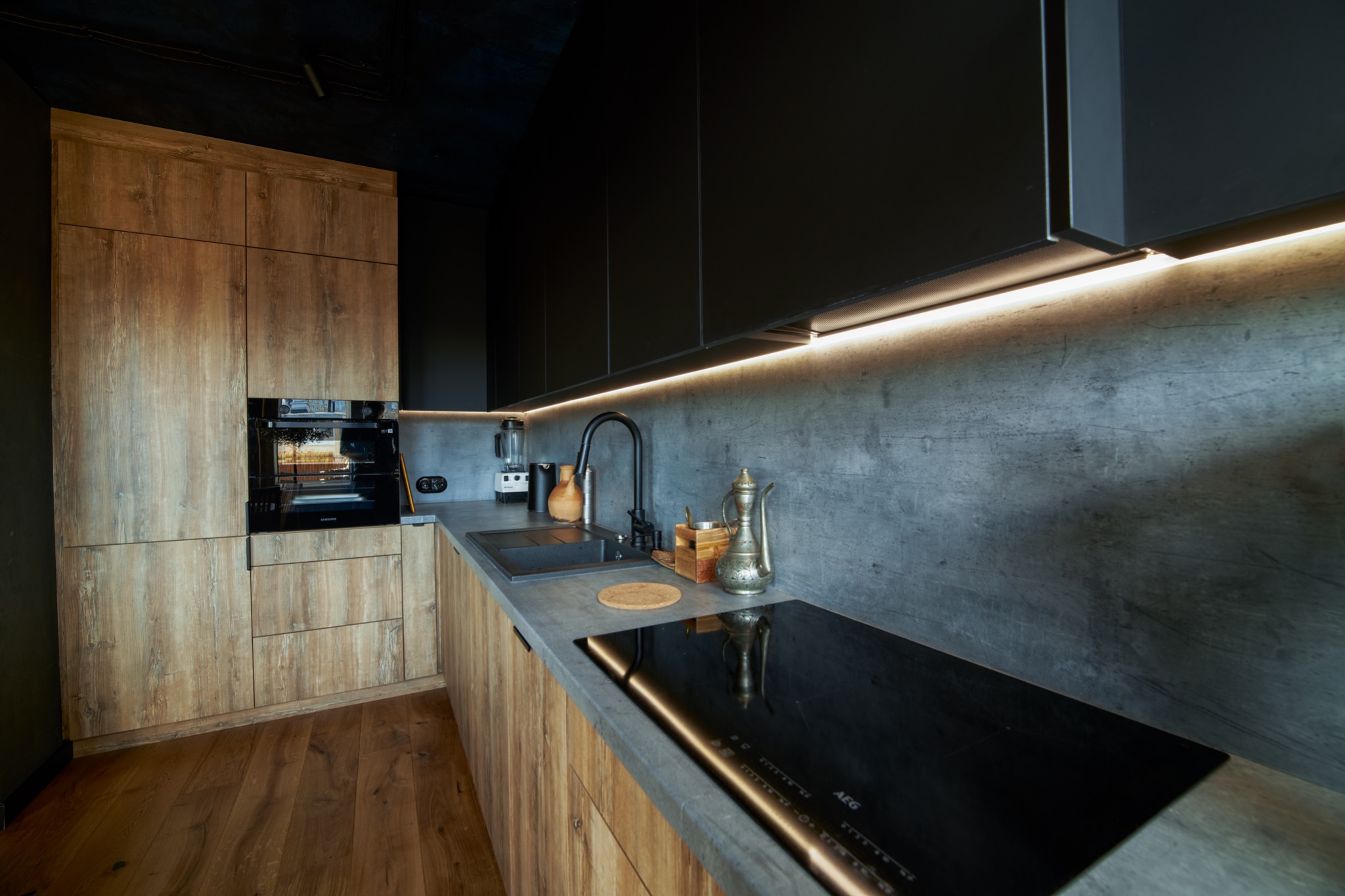 Nowoczesna kuchnia z drewnianymi frontami szafek i czarnymi elementami, oświetlenie LED podszafkowe, betonowa ściana, płyta indukcyjna AEG.
