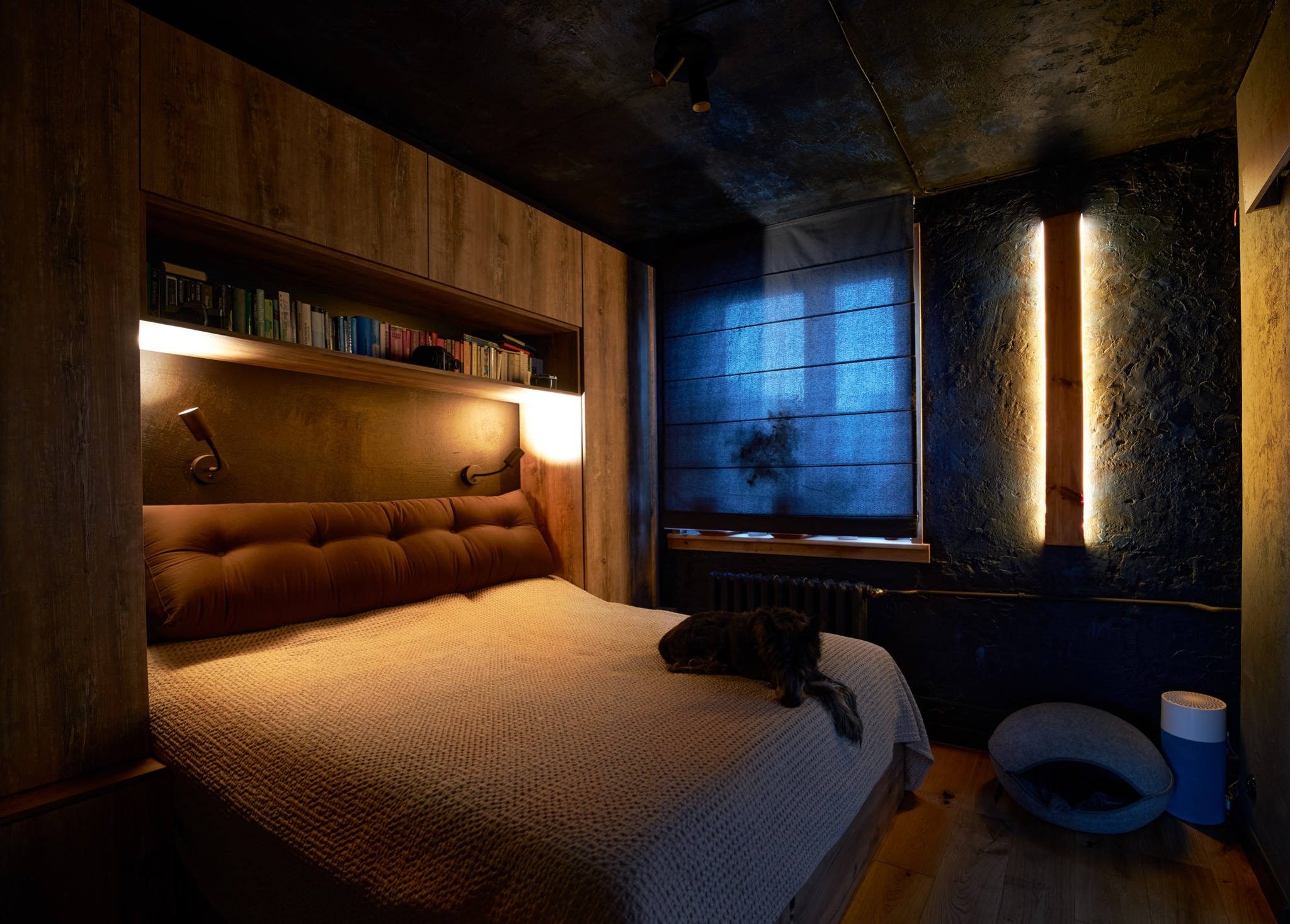 Nowoczesna sypialnia z łóżkiem w zabudowie z drewnianymi szafkami, półką na książki i oświetleniem punktowym, ciemna ściana z pionowym oświetleniem LED, rolety rzymskie w oknie, na łóżku leży pies,...