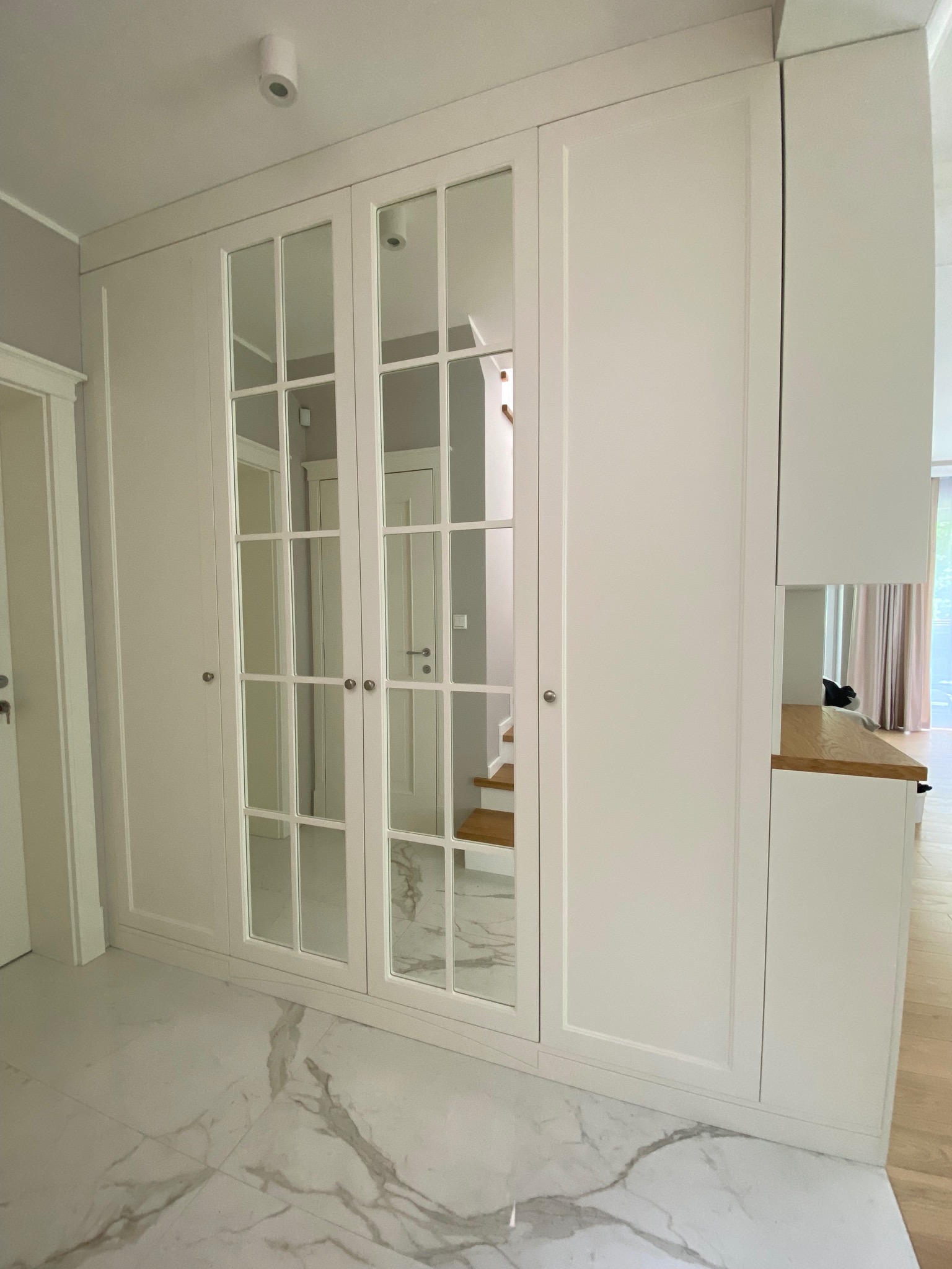 Biała szafa wnękowa z lustrzanymi drzwiami w stylu angielskim, odbicie schodów w lustrach, marmurowa podłoga z szarymi żyłami, minimalistyczny design.