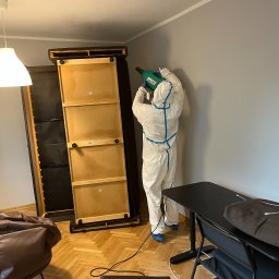 DEZPRO - Dezynsekcja Dezynfekcja Deratyzacja | Odpluskwianie Warszawa - Osoba w kombinezonie ochronnym z niebieskimi paskami, niebieskich ochraniaczach na buty i rękawicach, rozpyla środek z zielonego opryskiwacza na ścianę w pokoju z meblami.