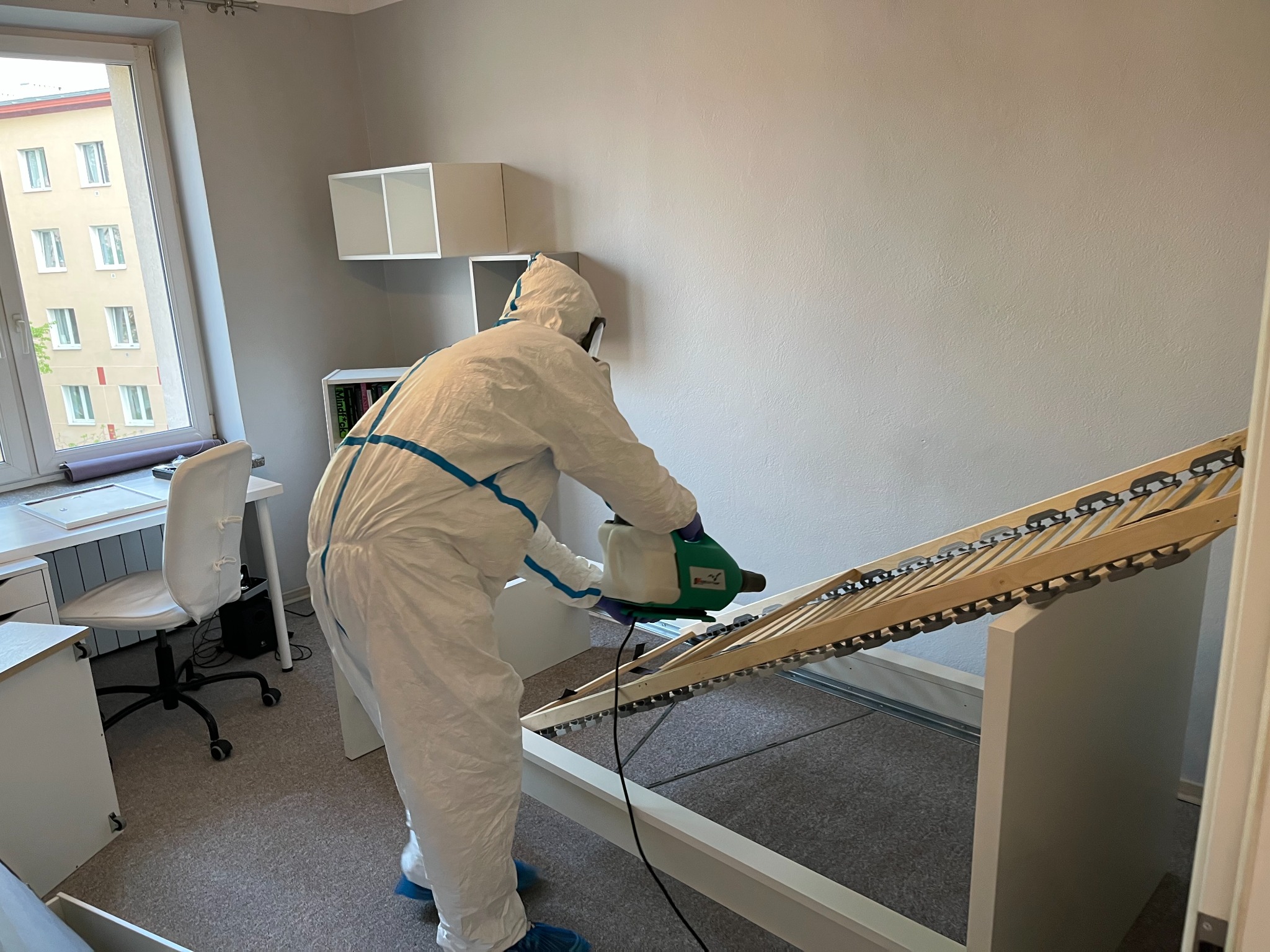 Osoba w kombinezonie ochronnym, z niebieskimi akcentami, rozpyla środek z zielonego opryskiwacza na stelaż łóżka w jasnym pokoju.