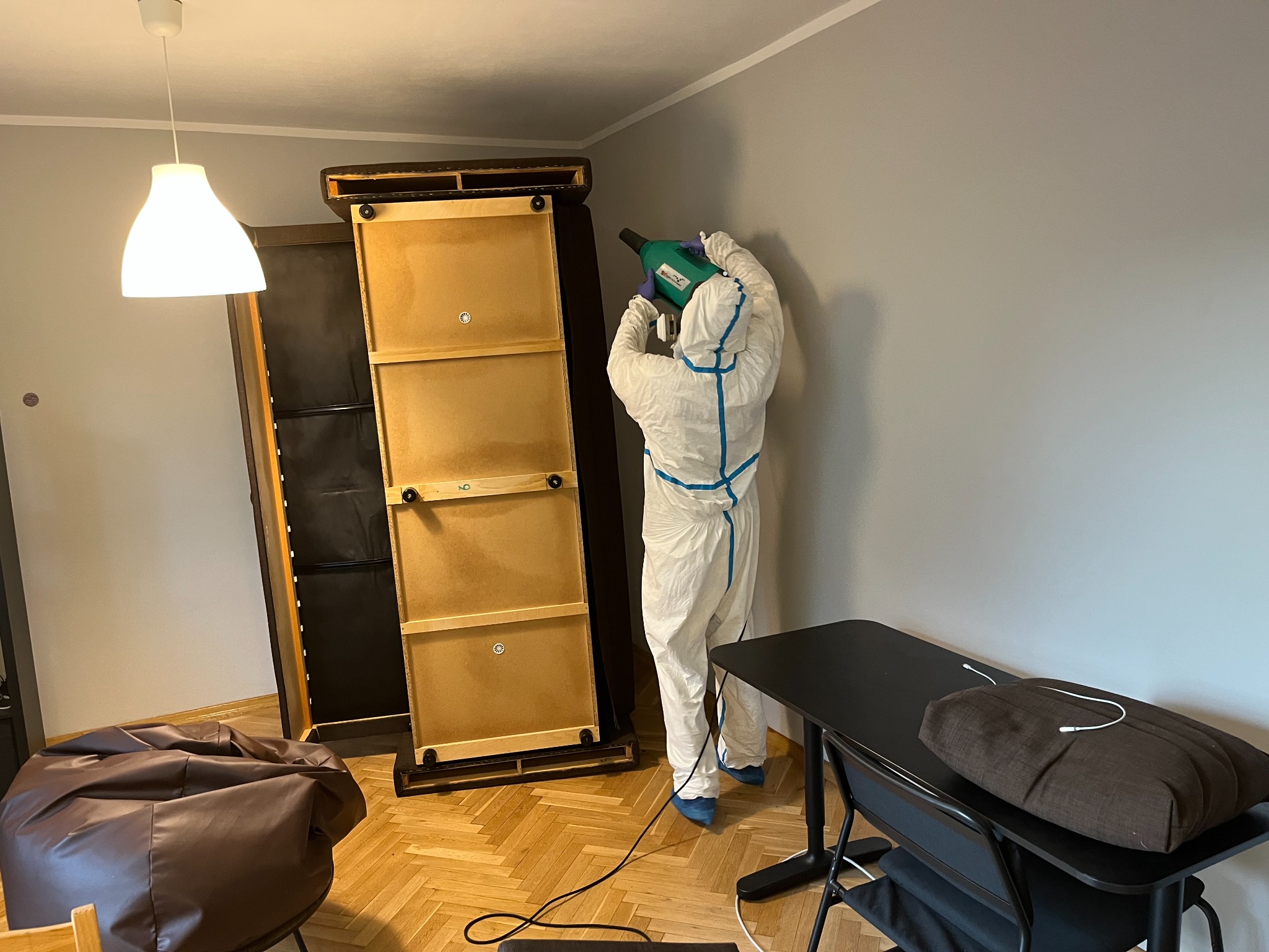 Osoba w kombinezonie ochronnym z niebieskimi paskami, niebieskich ochraniaczach na buty i rękawicach, rozpyla środek z zielonego opryskiwacza na ścianę w pokoju z meblami.