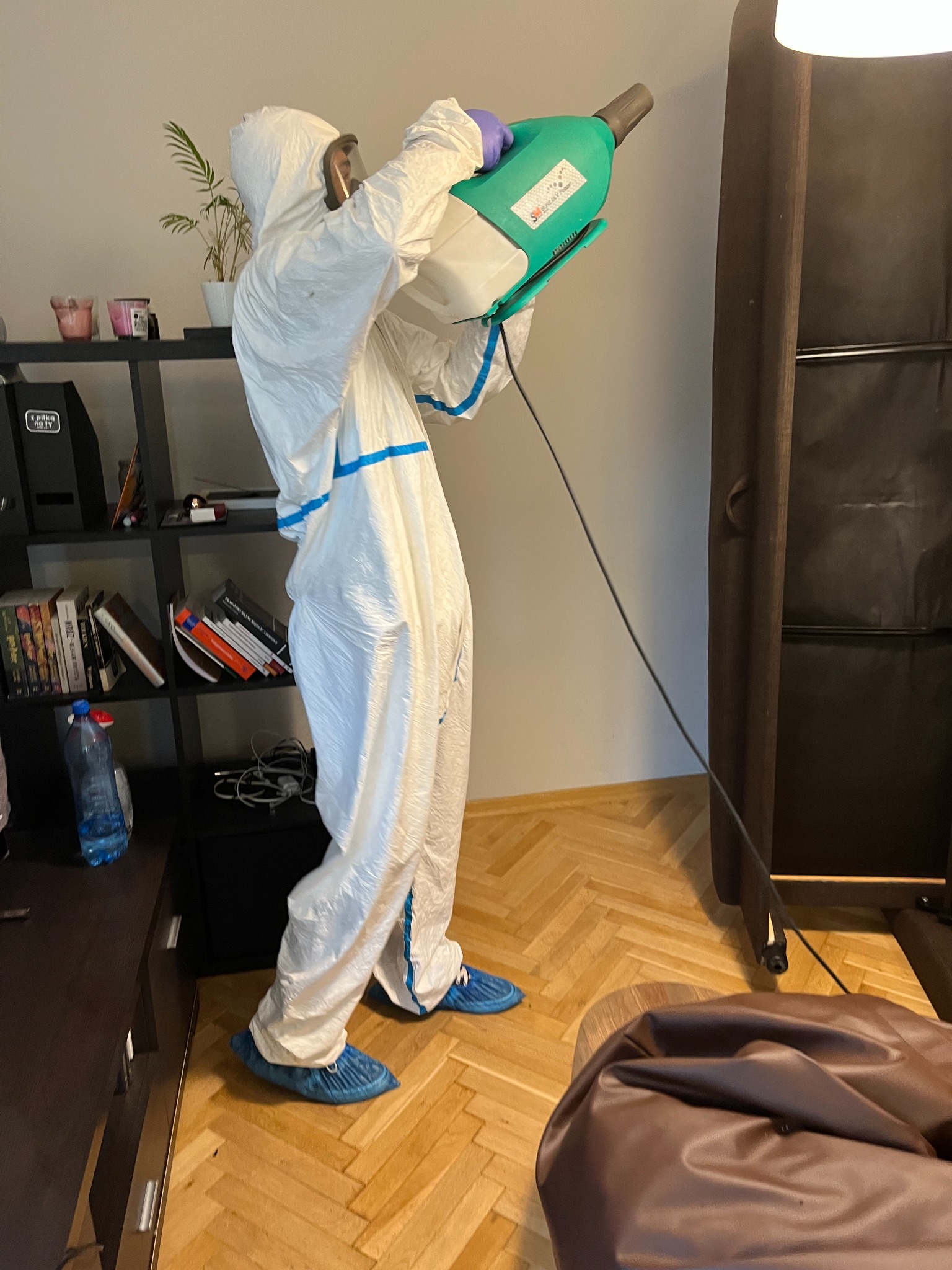 Osoba w kombinezonie ochronnym i niebieskich ochraniaczach na buty, używająca zielonego opryskiwacza w pomieszczeniu z drewnianą podłogą i regałem z książkami.