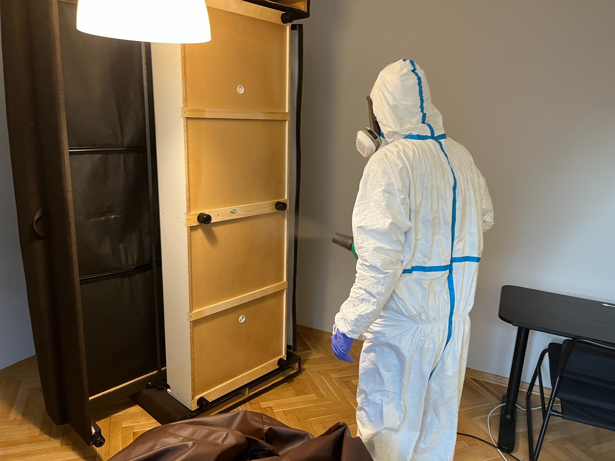 Osoba w kombinezonie ochronnym i masce rozpyla środek na drewnianą konstrukcję z czterema panelami w pomieszczeniu z parkietem i czarnym biurkiem.