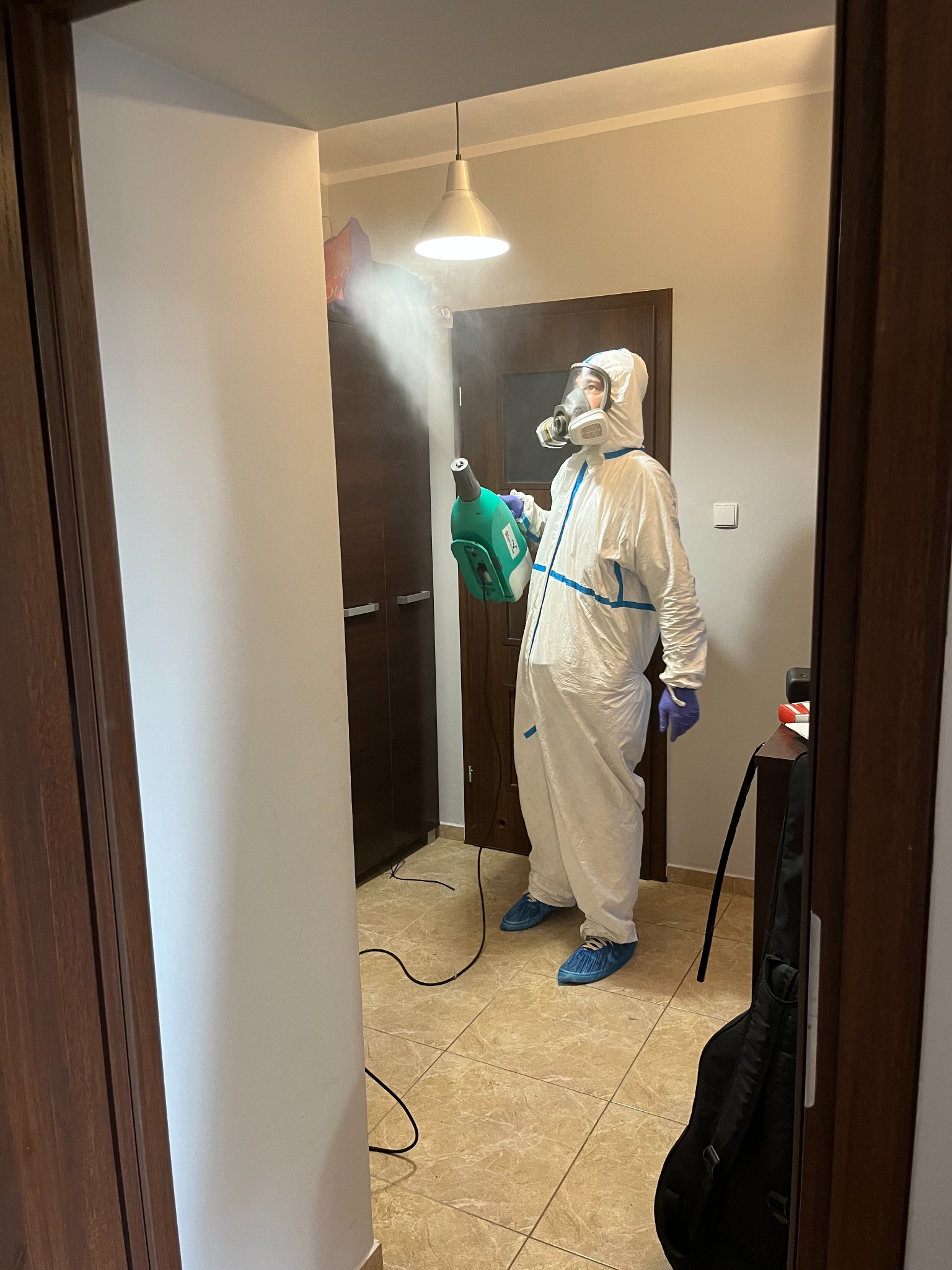 Osoba w kombinezonie ochronnym i masce rozpyla środek dezynfekujący w pomieszczeniu, widoczna mgiełka, podłoga wyłożona kafelkami.
