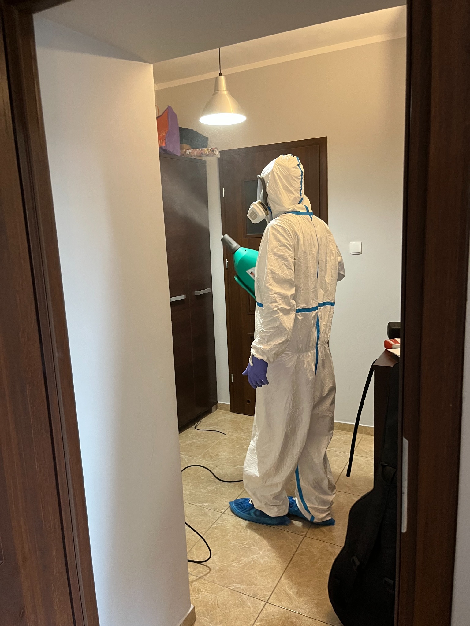 Osoba w kombinezonie ochronnym, masce i rękawicach, rozpyla środek z zielonego opryskiwacza w pomieszczeniu z szafą i lampą wiszącą.