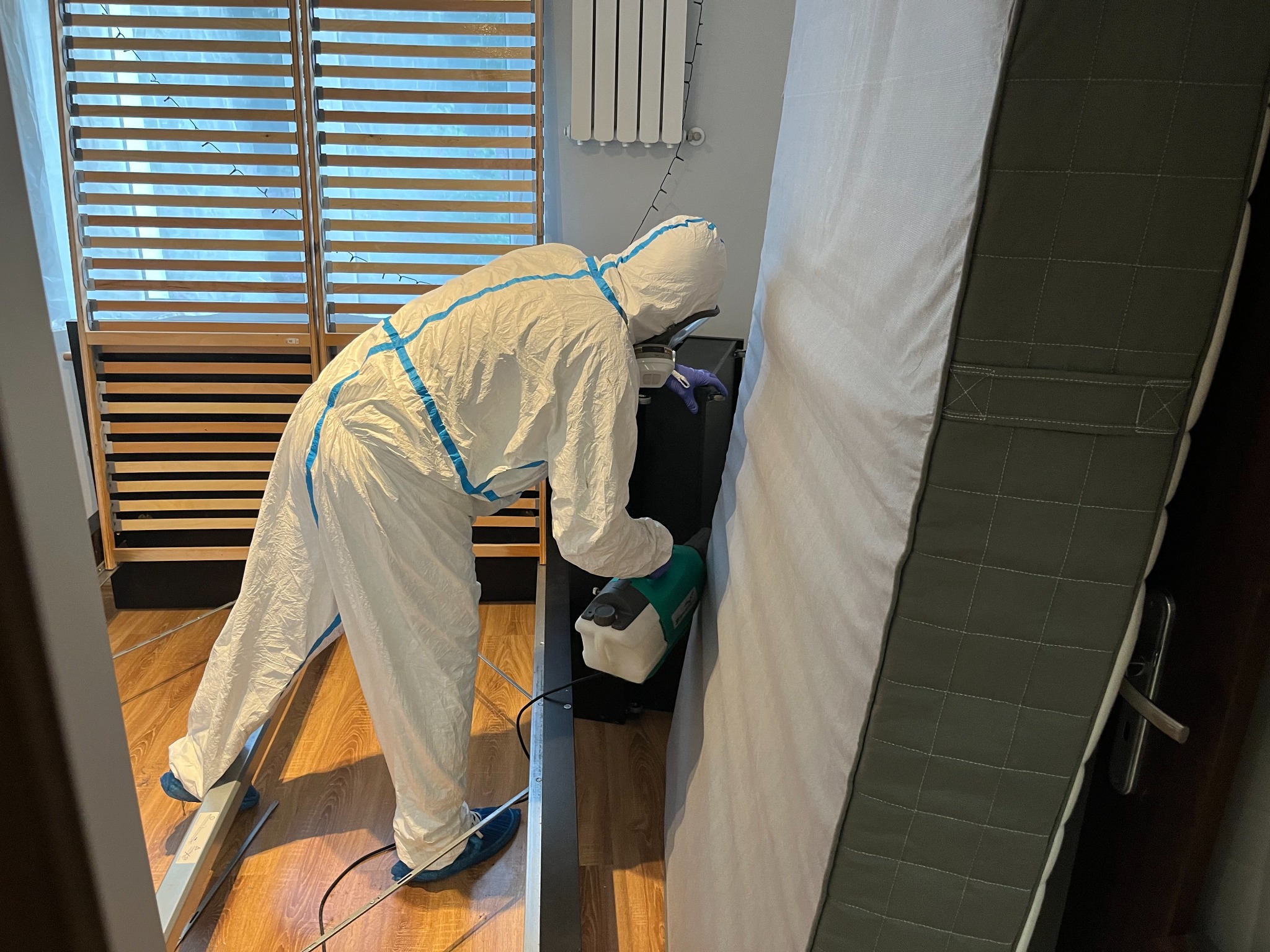 Osoba w kombinezonie ochronnym dezynfekuje ramę łóżka za pomocą opryskiwacza.