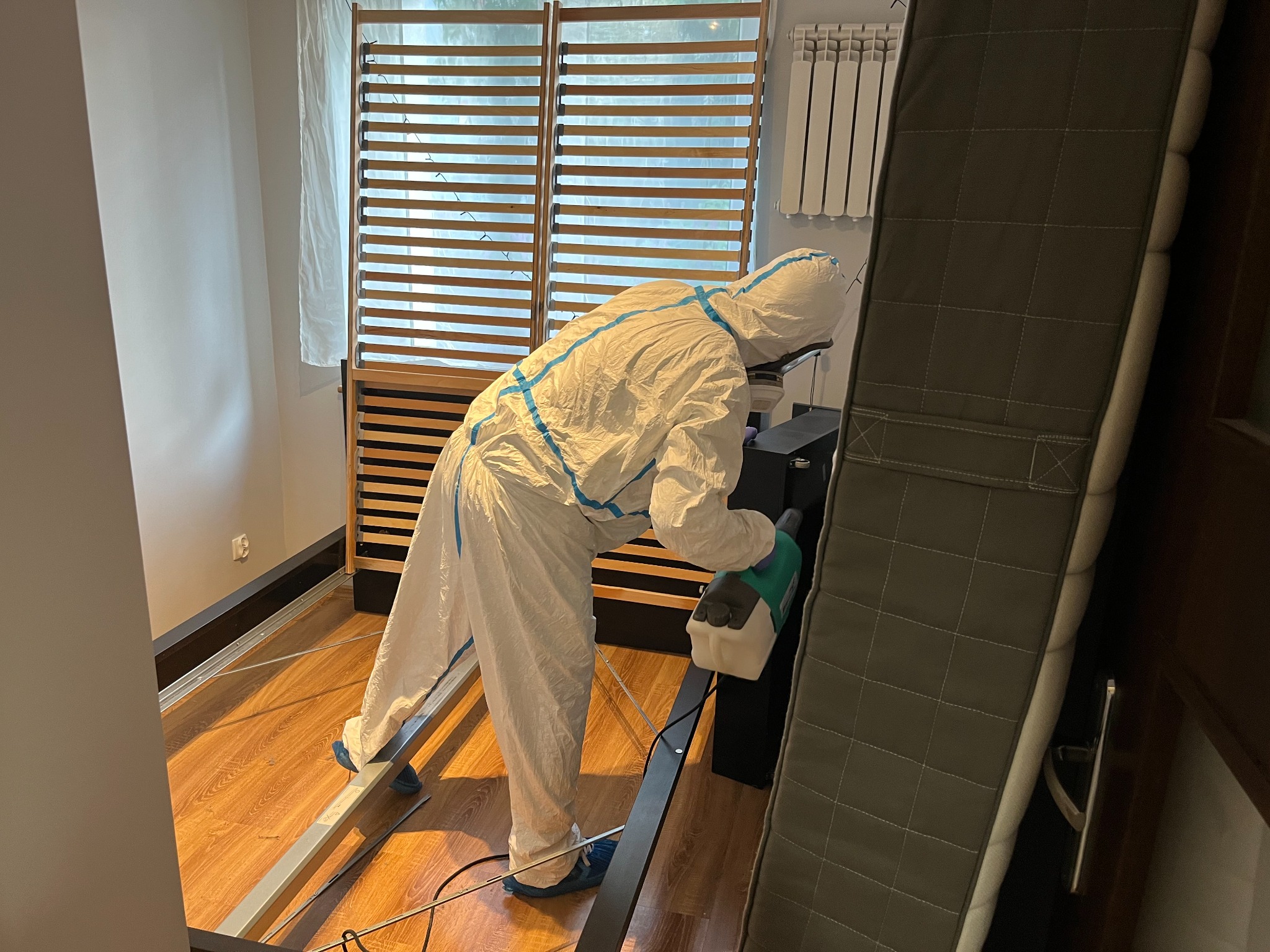 Osoba w kombinezonie ochronnym, z niebieskimi akcentami, opryskuje ramę łóżka za pomocą ręcznego opryskiwacza w pomieszczeniu z drewnianą podłogą.