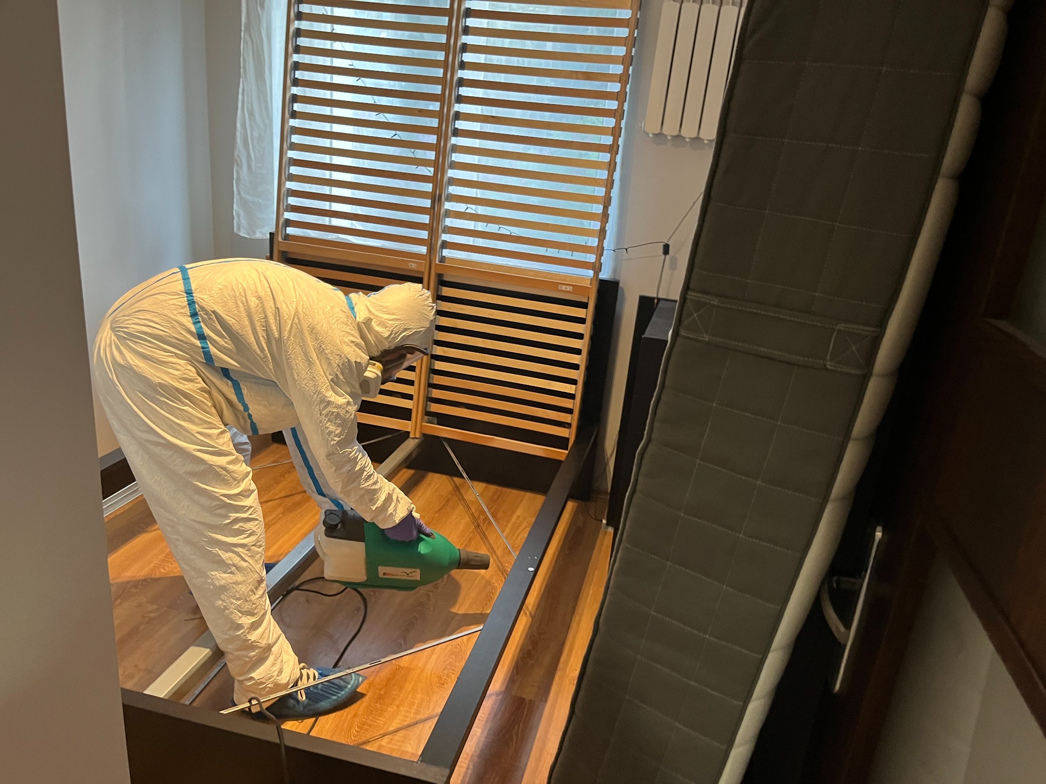 Osoba w kombinezonie ochronnym, rękawicach i niebieskich ochraniaczach na obuwie, używa zielonego opryskiwacza w pomieszczeniu z rozłożonym łóżkiem.