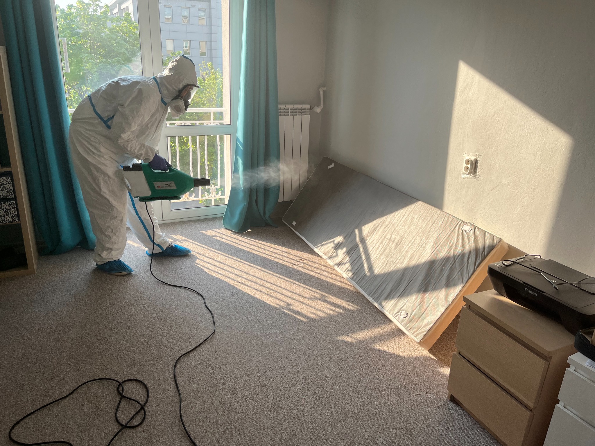 Osoba w kombinezonie ochronnym i masce rozpyla preparat za pomocą zamgławiacza w jasnym pokoju z łóżkiem opartym o ścianę, widoczne promienie słoneczne wpadające przez okno.