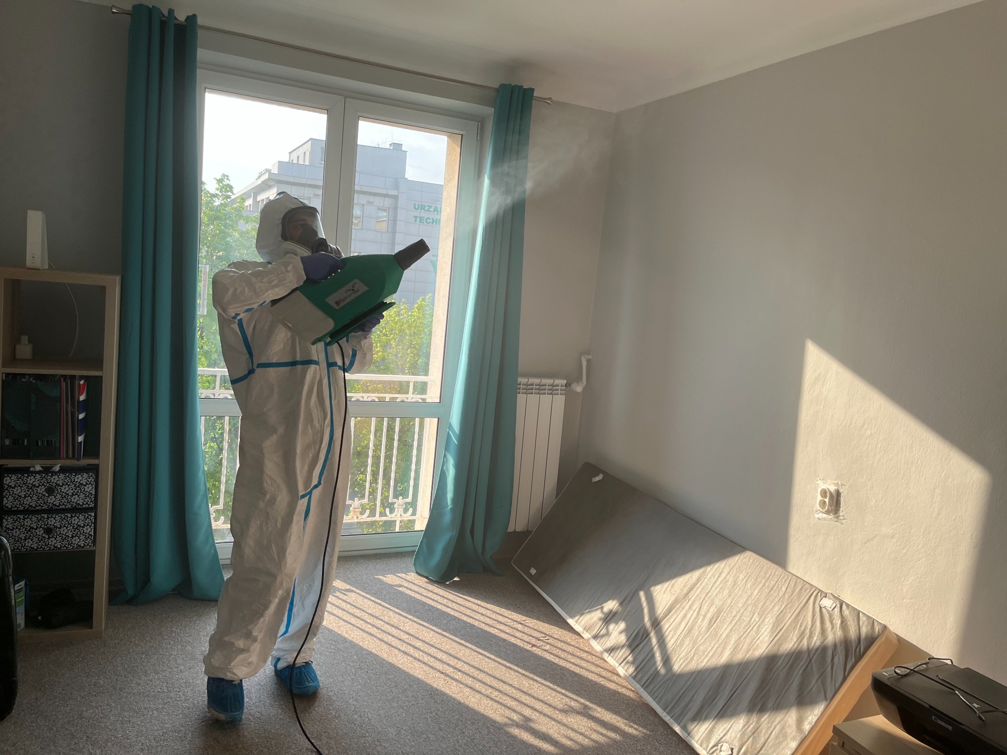Pracownik w kombinezonie ochronnym, masce i niebieskich ochraniaczach na obuwiu, rozpyla preparat w pomieszczeniu z oknem i złożonym materacem.