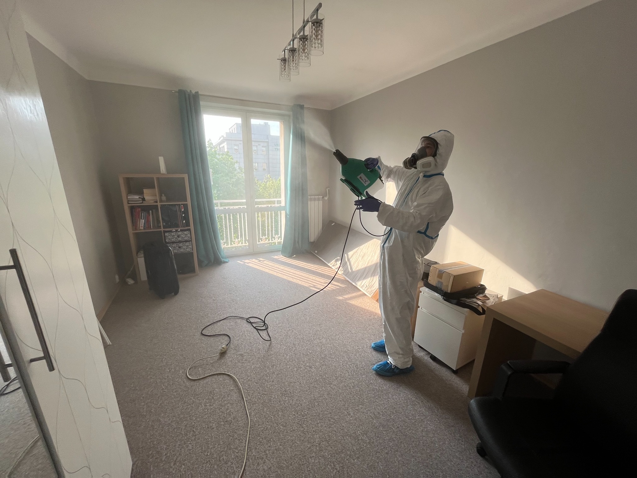 Pracownik w kombinezonie ochronnym i masce rozpyla preparat w pomieszczeniu mieszkalnym, widoczny sprzęt do oprysku i kabel zasilający na podłodze.