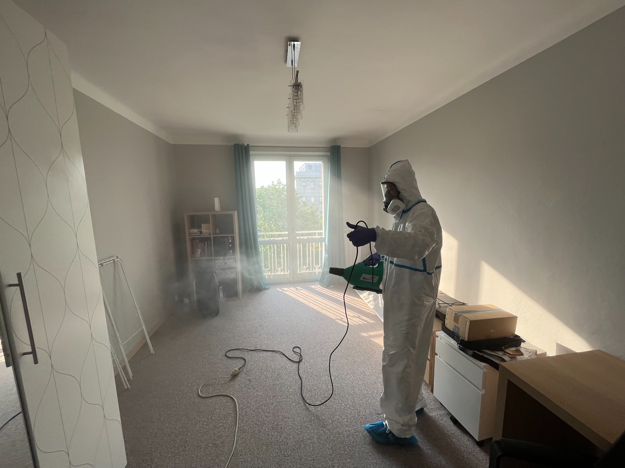 Osoba w kombinezonie ochronnym i masce rozpyla środek z elektrycznego opryskiwacza w jasnym pokoju z balkonem, widoczna mgiełka w powietrzu.