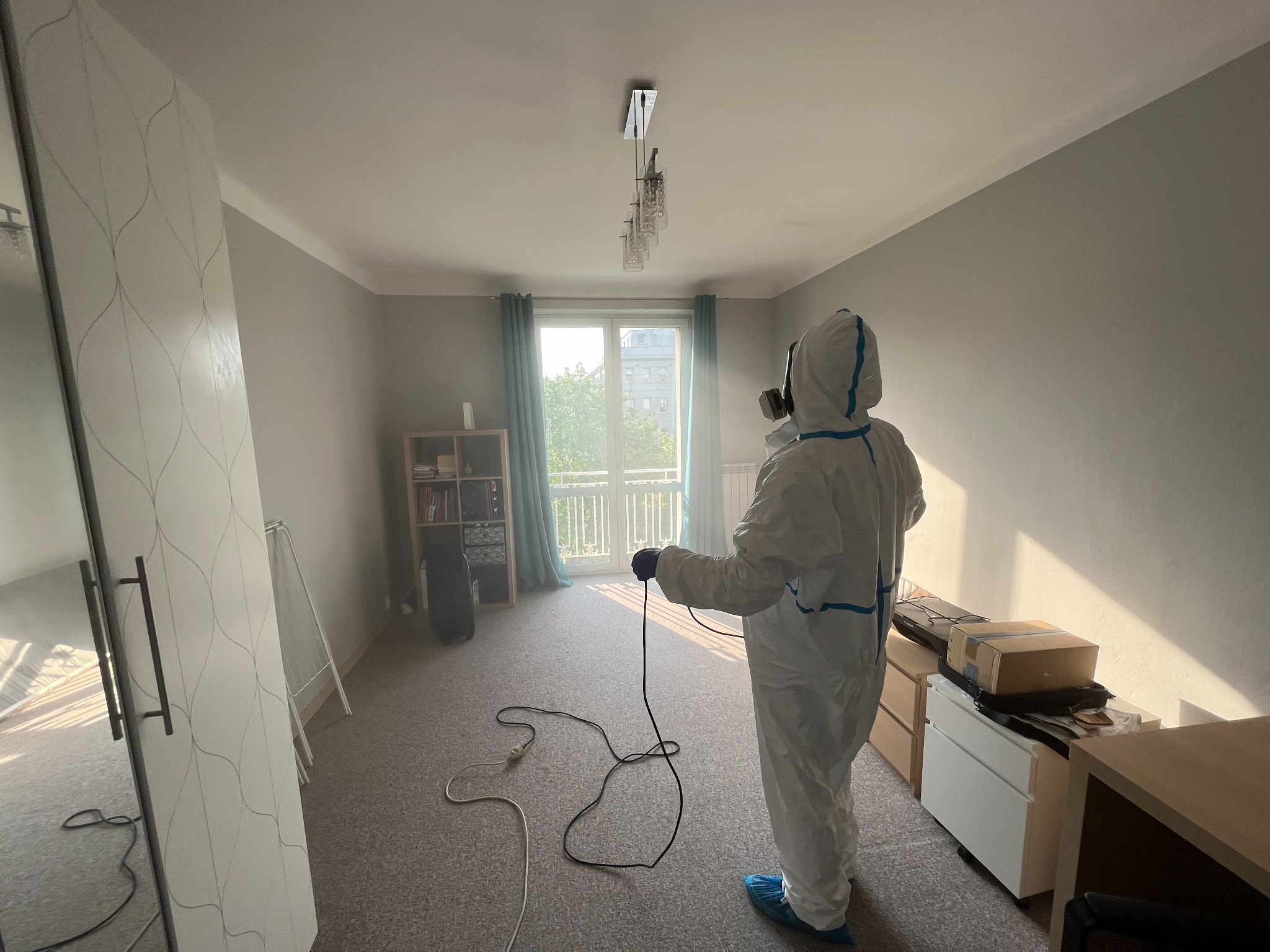 Osoba w kombinezonie ochronnym i masce gazowej, trzymająca w ręku przewód urządzenia do dezynsekcji w jasnym pokoju z dywanem i meblami, przygotowuje się do zabiegu.