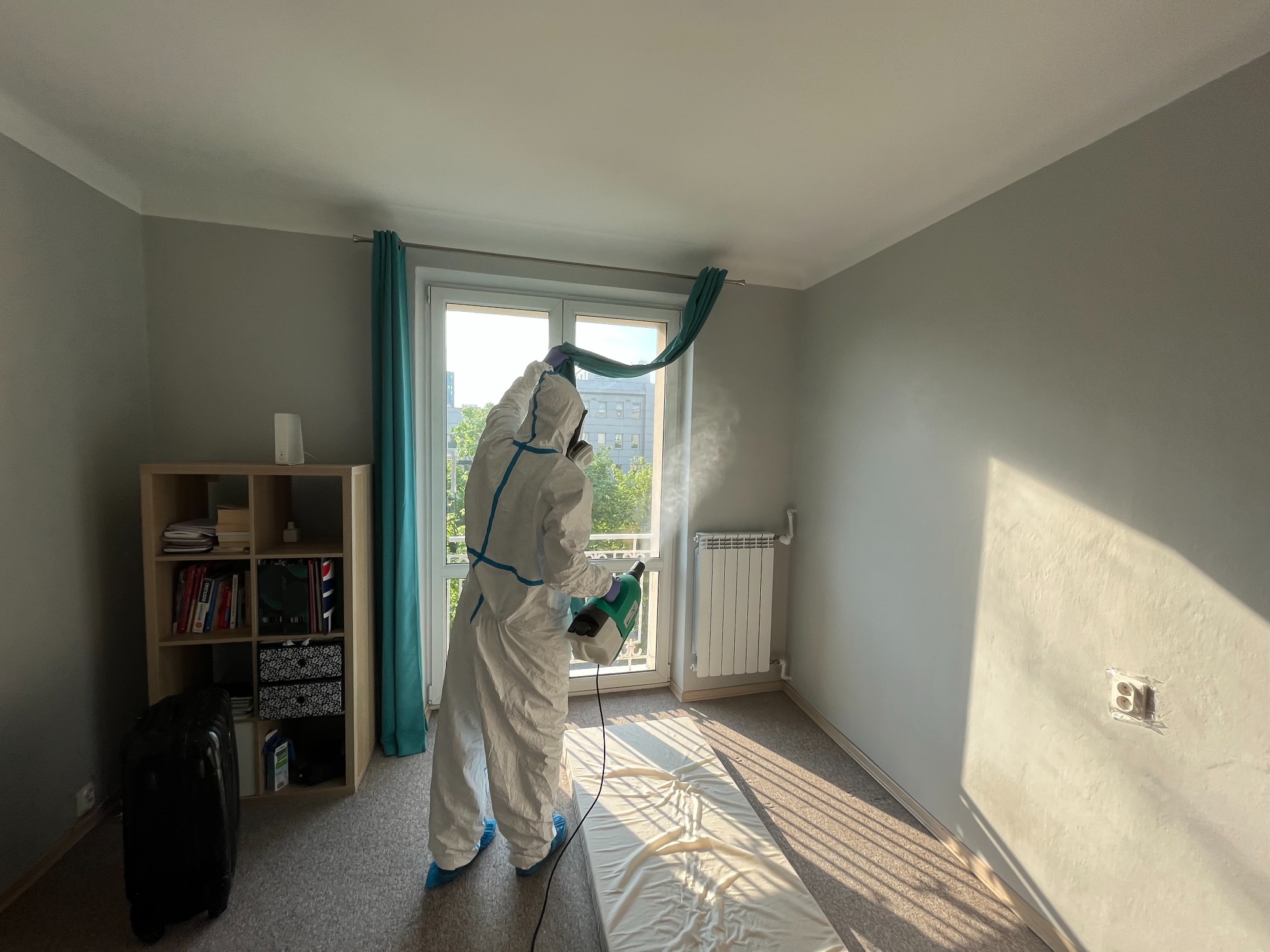 Osoba w kombinezonie ochronnym spryskuje preparatem sufit w pokoju z zasłoniętym oknem, widoczny szafka z książkami i walizka.