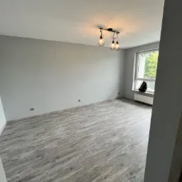 Pusty pokój po remoncie z szarymi ścianami, panelami podłogowymi w odcieniach szarości i nowoczesną lampą sufitową z trzema żarówkami w stylu industrialnym.