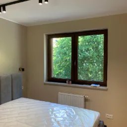 Nowoczesna sypialnia z szarym łóżkiem z zagłówkiem, oświetleniem szynowym na suficie i widokiem na kwitnące drzewo zza okna w brązowej ramie.