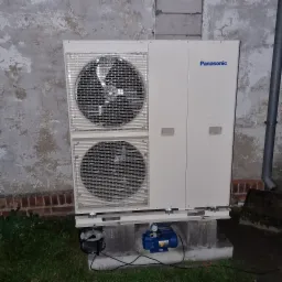 Dwu-wentylatorowa jednostka zewnętrzna pompy ciepła Panasonic, zamontowana na betonowym fundamencie z widoczną niebieską pompą próżniową i przedłużaczem elektrycznym, ustawiona na tle ściany...