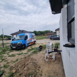 Term Instal Technika Grzewcza Sebastian Staszewski - Zewnętrzna jednostka klimatyzacji zamontowana przy ścianie domu z widocznym firmowym vanem w tle, narzędzia i materiały rozłożone na ziemi.