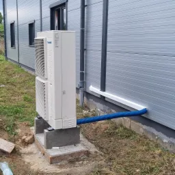 Zewnętrzna jednostka klimatyzacji Panasonic na betonowym fundamencie przy szarej ścianie budynku przemysłowego, z niebieskim wężem odprowadzającym skropliny.