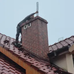 Instalator na dachu, montujący lub sprawdzający komin wentylacyjny, z widocznymi drabinkami dachowymi i ceglanym kominem.