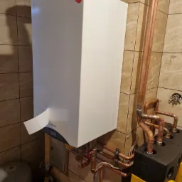 Biały, wiszący kocioł gazowy marki Termet z naklejką 'Polski Producent', podłączony do miedzianych rur instalacji grzewczej na tle beżowych płytek.