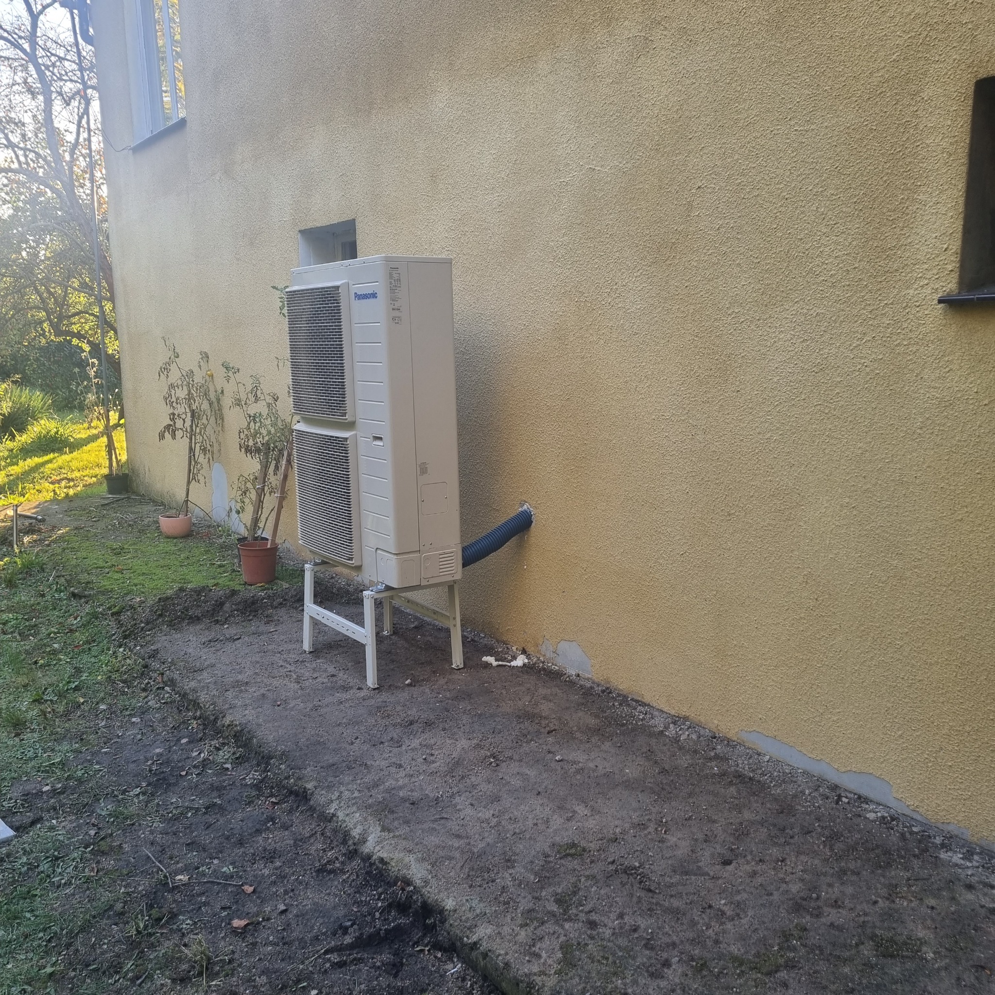 Dwuczęściowa jednostka zewnętrzna klimatyzacji marki Panasonic zamontowana na białym stelażu przy otynkowanej ścianie budynku; widoczny niebieski wąż odprowadzający skropliny.