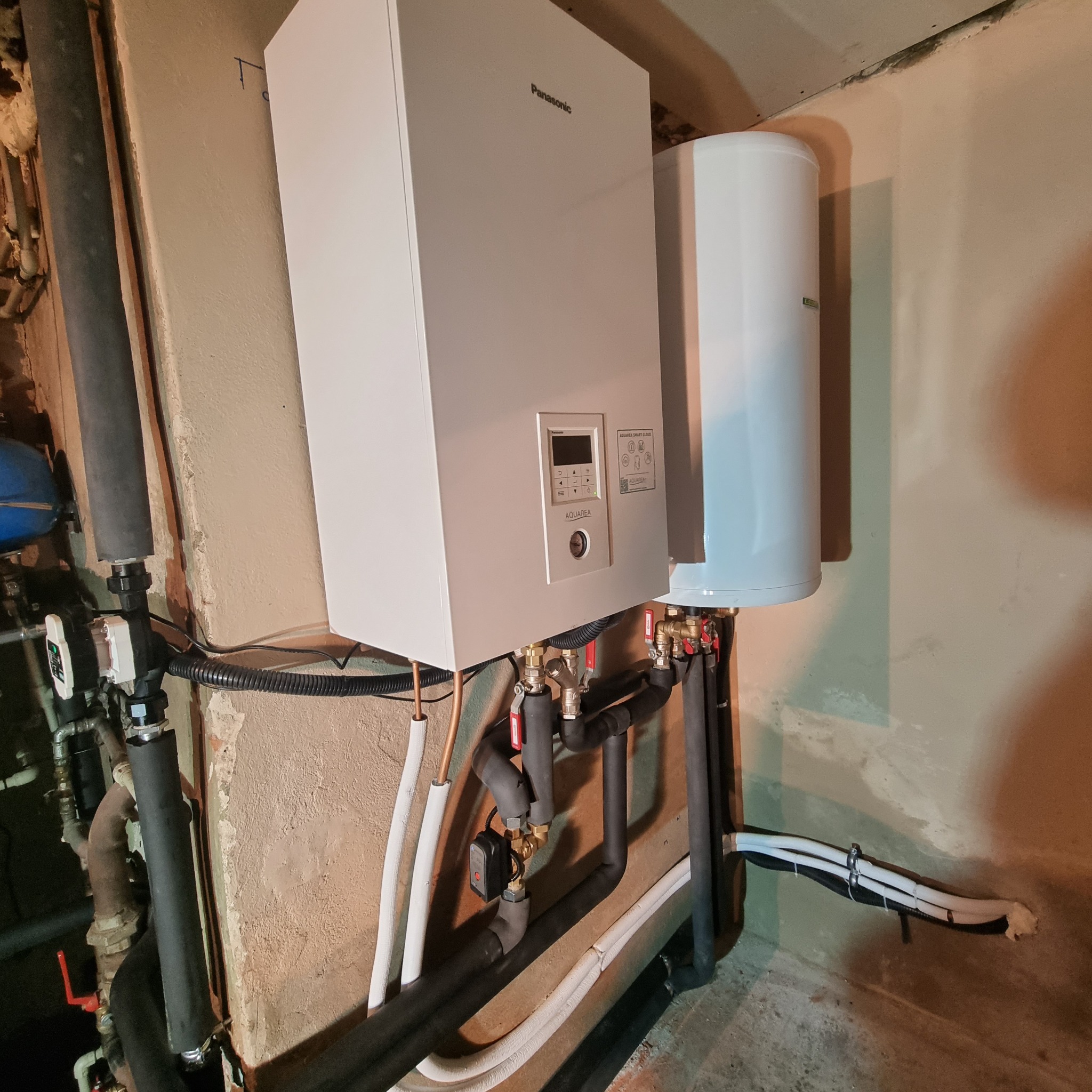 Biała jednostka wewnętrzna pompy ciepła Panasonic Aquarea Smart Cloud wisząca na ścianie, z widocznymi rurami miedzianymi i izolowanymi rurami czarnymi oraz zbiornikiem na wodę po prawej stronie.