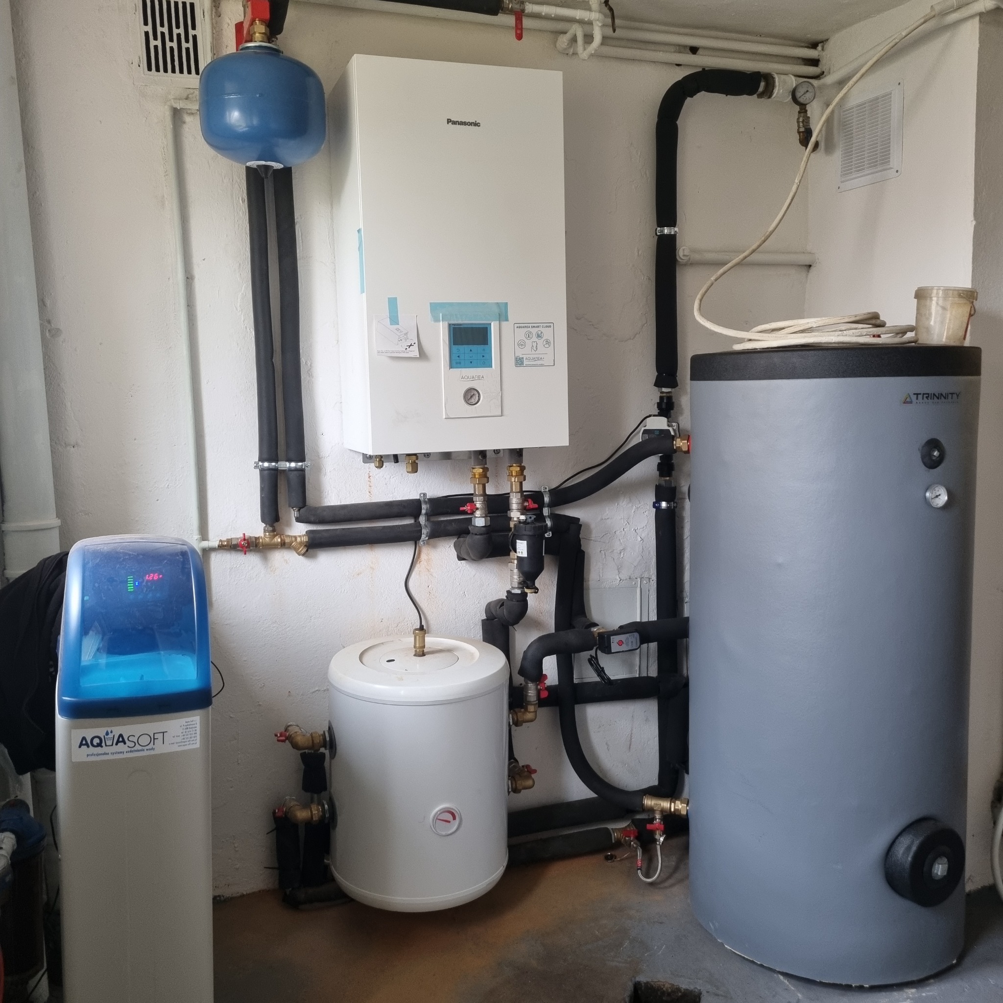 Instalacja grzewcza w kotłowni: biały piec gazowy Panasonic, szary zbiornik Trinity i niebieski zmiękczacz wody AquaSoft, połączone czarnymi rurami w izolacji, widoczne zawory i manometry.
