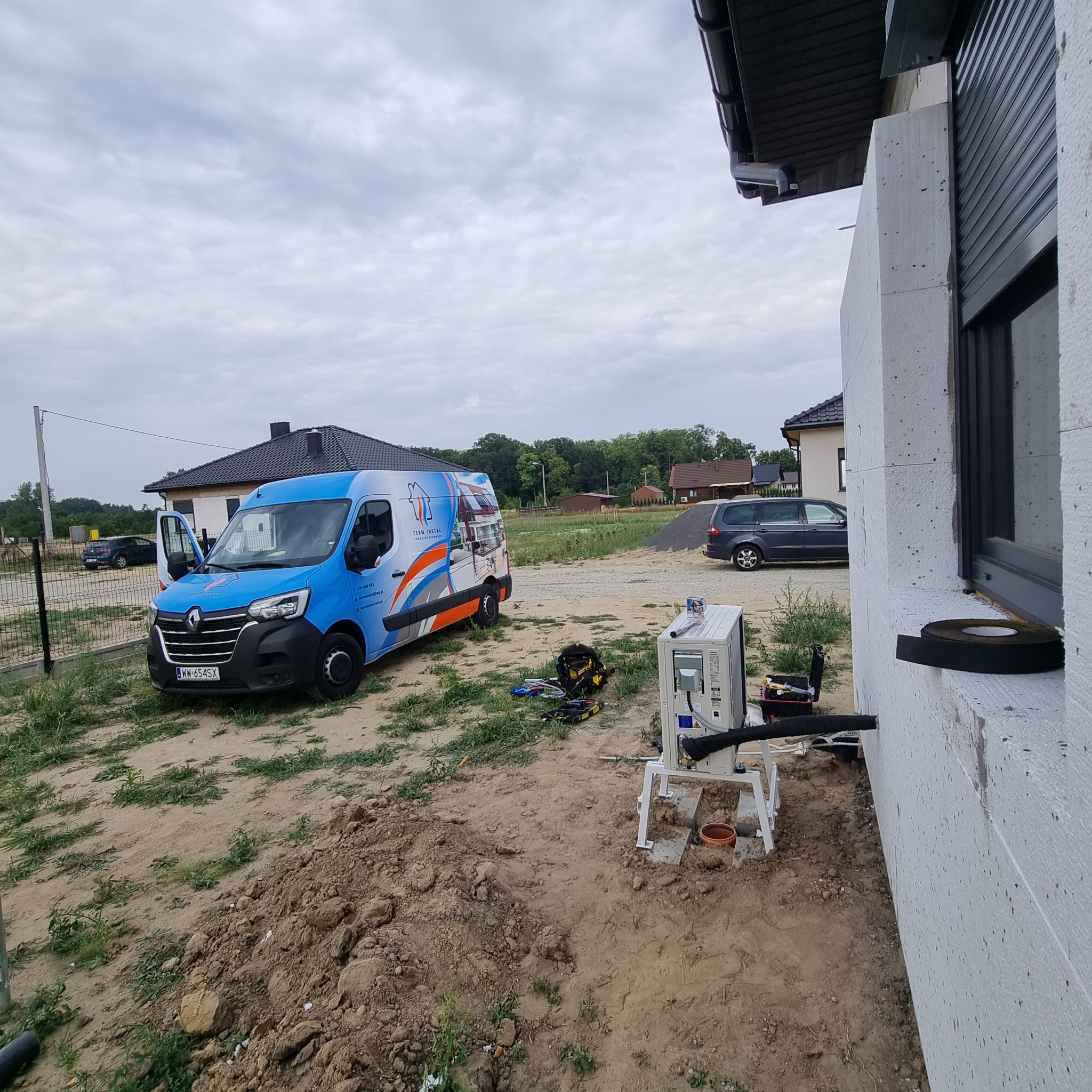 Zewnętrzna jednostka klimatyzacji zamontowana przy ścianie domu z widocznym firmowym vanem w tle, narzędzia i materiały rozłożone na ziemi.