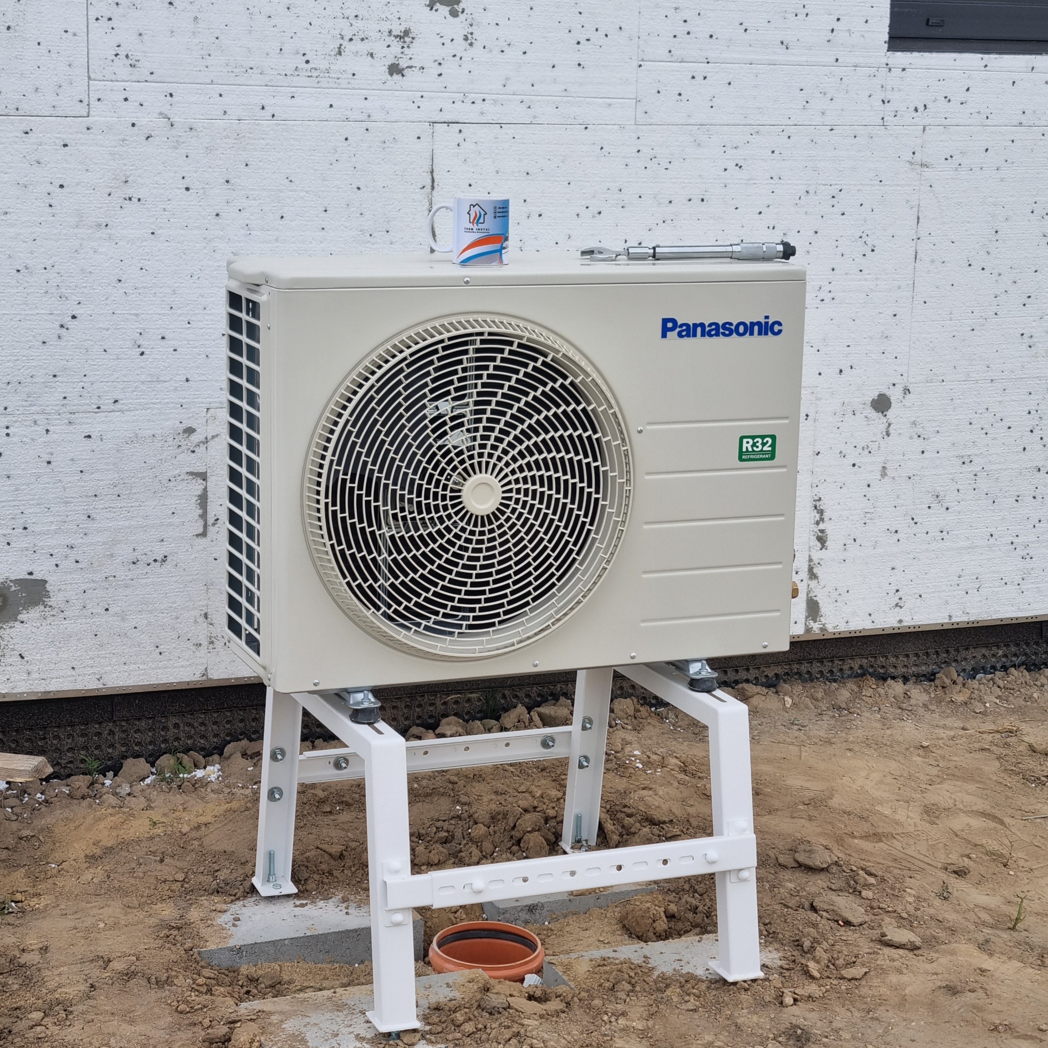 Zewnętrzna jednostka klimatyzacyjna Panasonic na białym, metalowym stelażu, z kubkiem firmowym i kluczem nastawnym na górze, stoi na ziemi przy ocieplonej ścianie budynku.