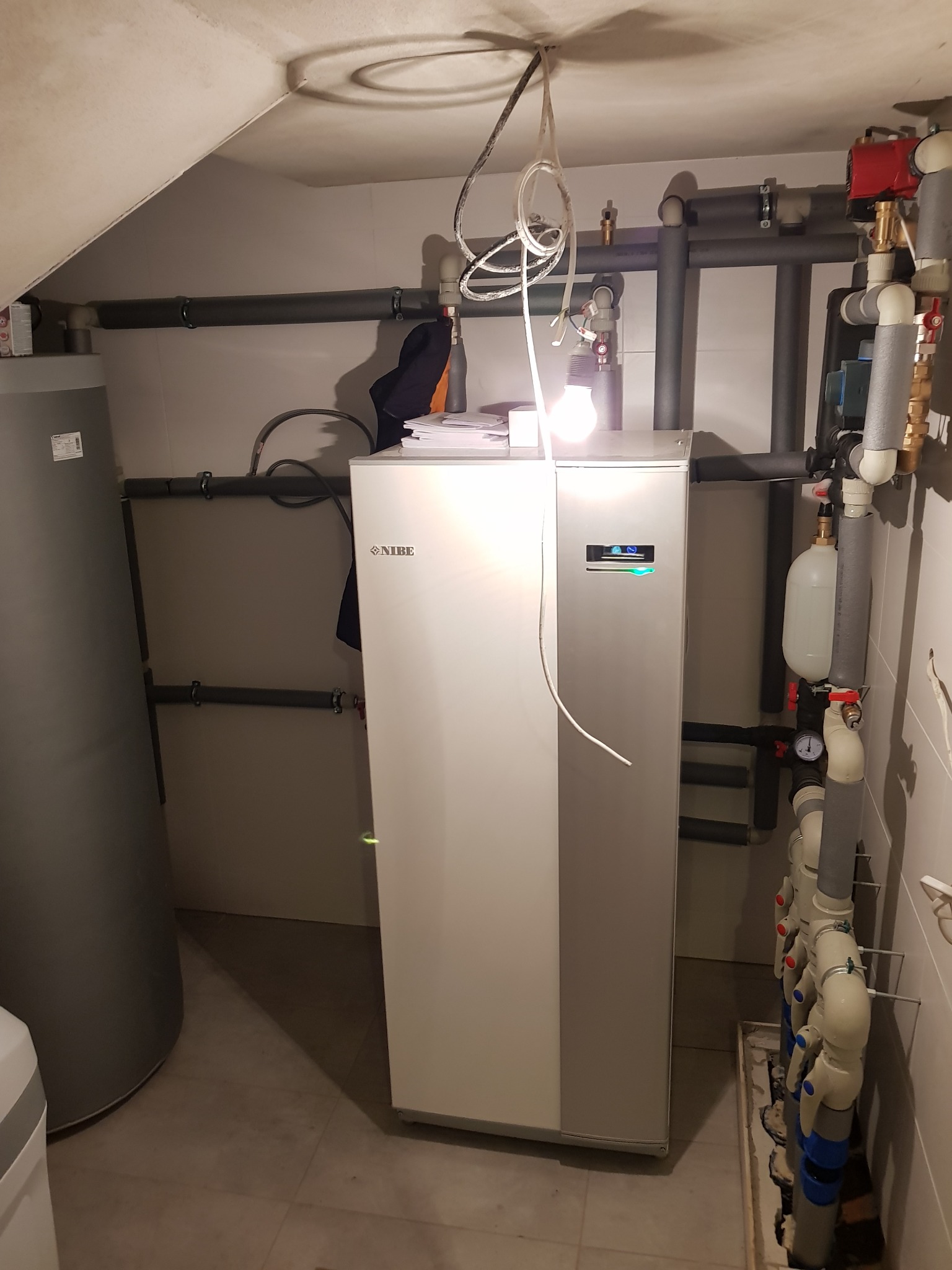 Nowoczesna instalacja grzewcza z szarą pompą ciepła Nibe, widoczne rury instalacyjne z izolacją, manometr i zawory w pomieszczeniu technicznym oświetlonym żarówką.