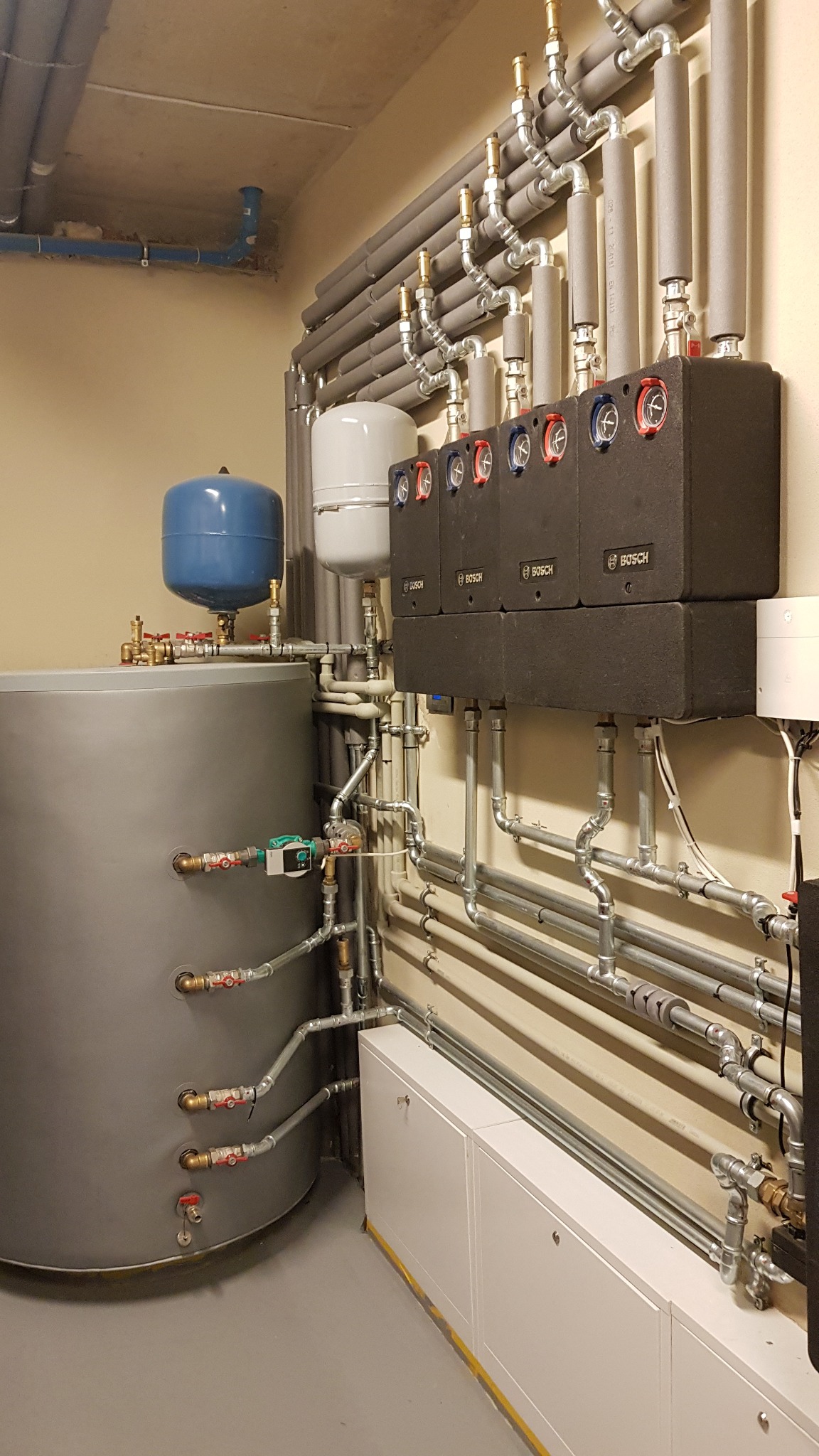 Kotłownia z szarym zbiornikiem akumulacyjnym, niebieskim naczyniem przeponowym, czterema czarnymi modułami Bosch z czerwono-niebieskimi regulatorami, oraz licznymi rurami z izolacją termiczną.