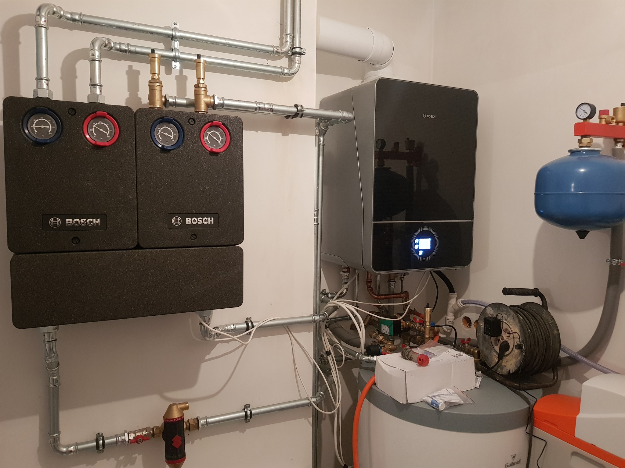 Zainstalowany system grzewczy Bosch z dwoma panelami sterowania, kotłem kondensacyjnym, zbiornikiem i kompletną instalacją hydrauliczną z rurami mocowanymi do ściany.