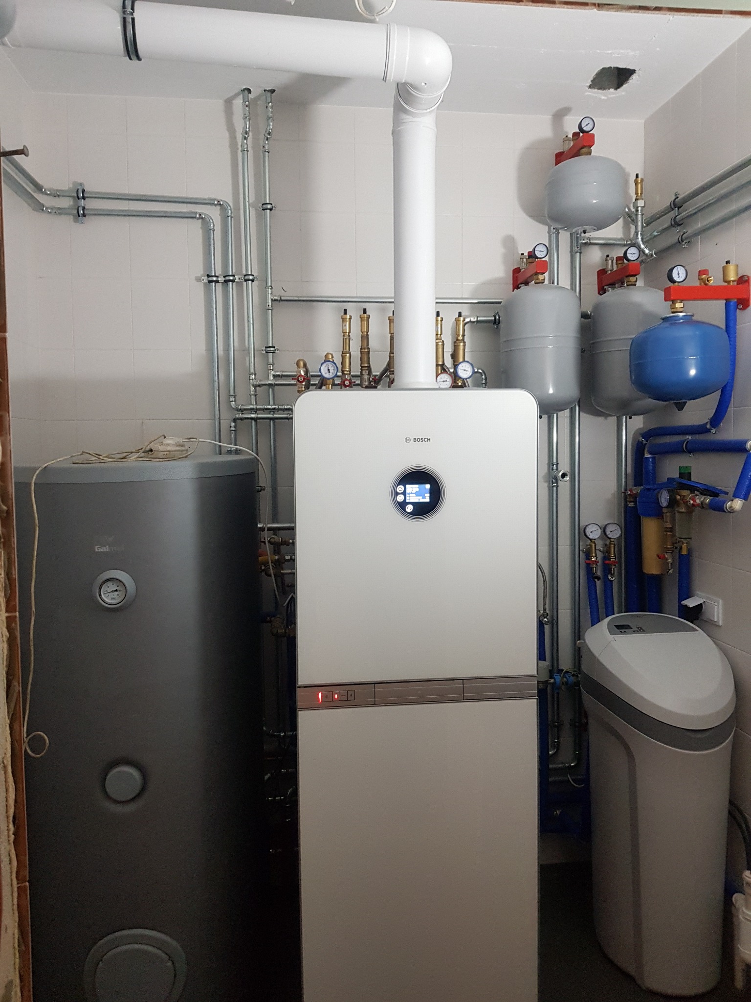 Kotłownia z nowoczesnym kotłem gazowym Bosch z okrągłym wyświetlaczem, zbiornikiem na wodę, zbiornikami wyrównawczymi i zmiękczaczem wody, instalacja hydrauliczna z rur miedzianych i plastikowych.